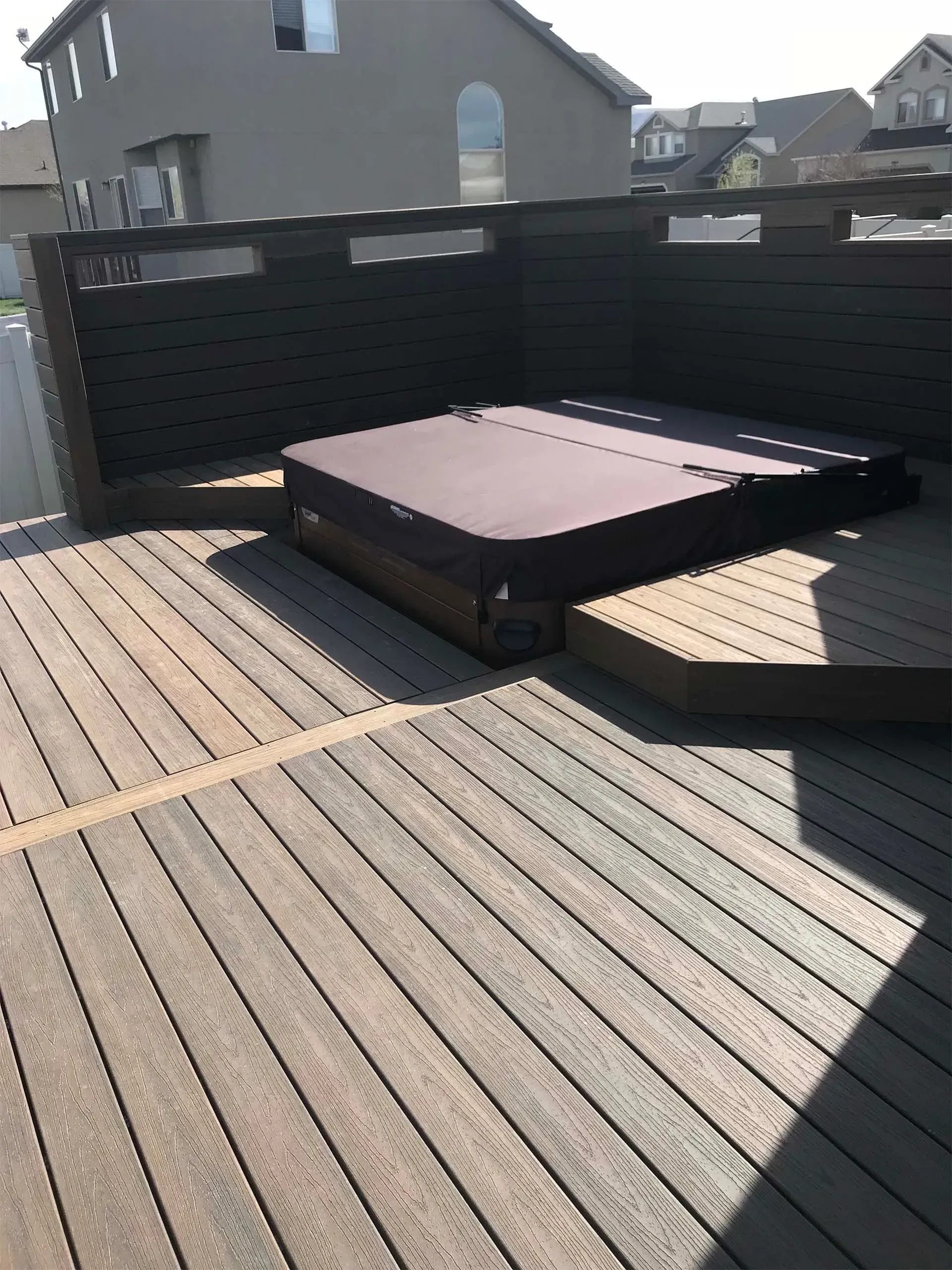 Hot tub with cover on a wooden deck, surrounded by a dark fence, in a sunny backyard.