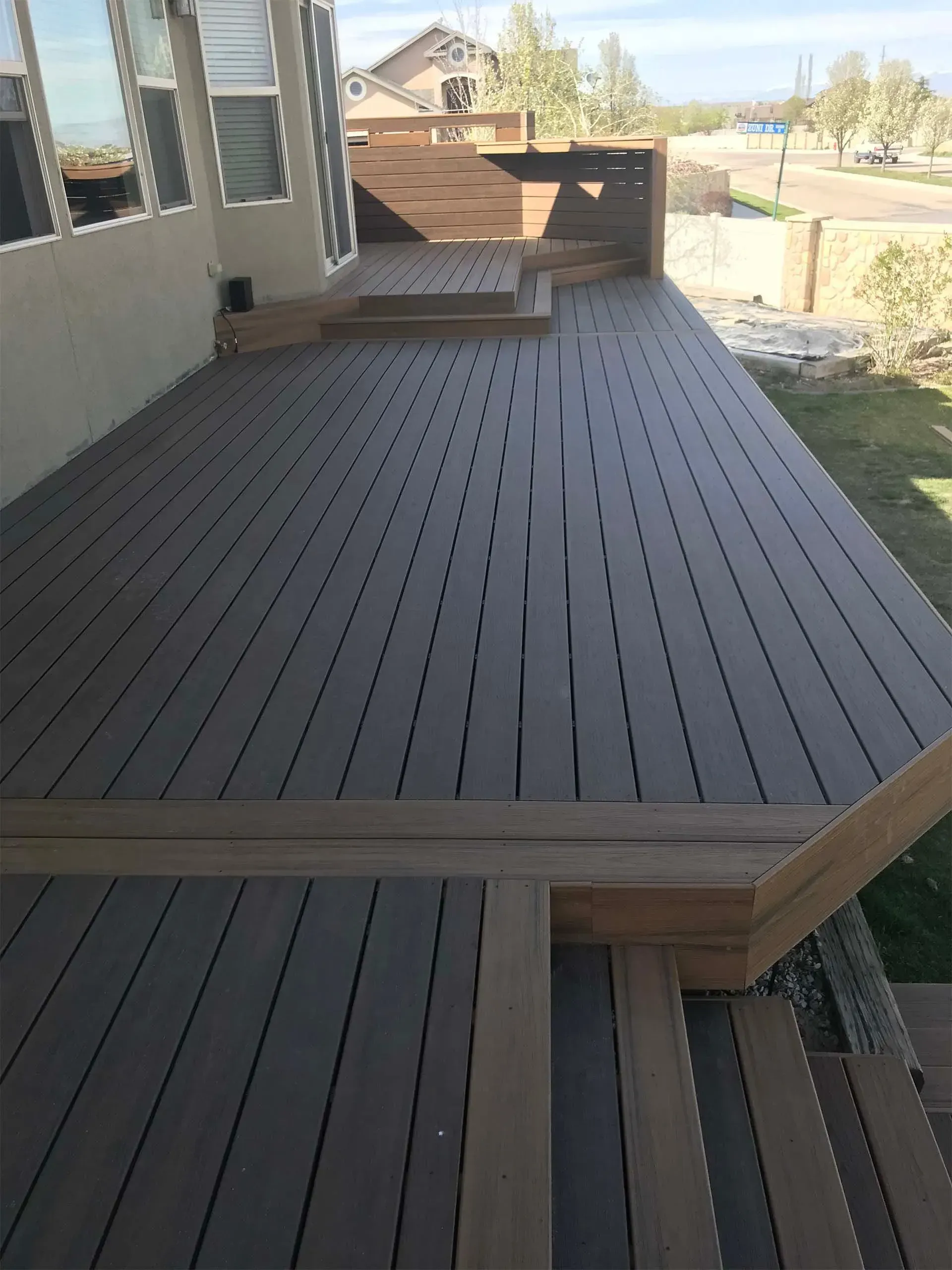 A multi-level composite deck with steps, in shades of gray and brown, next to a house with windows.