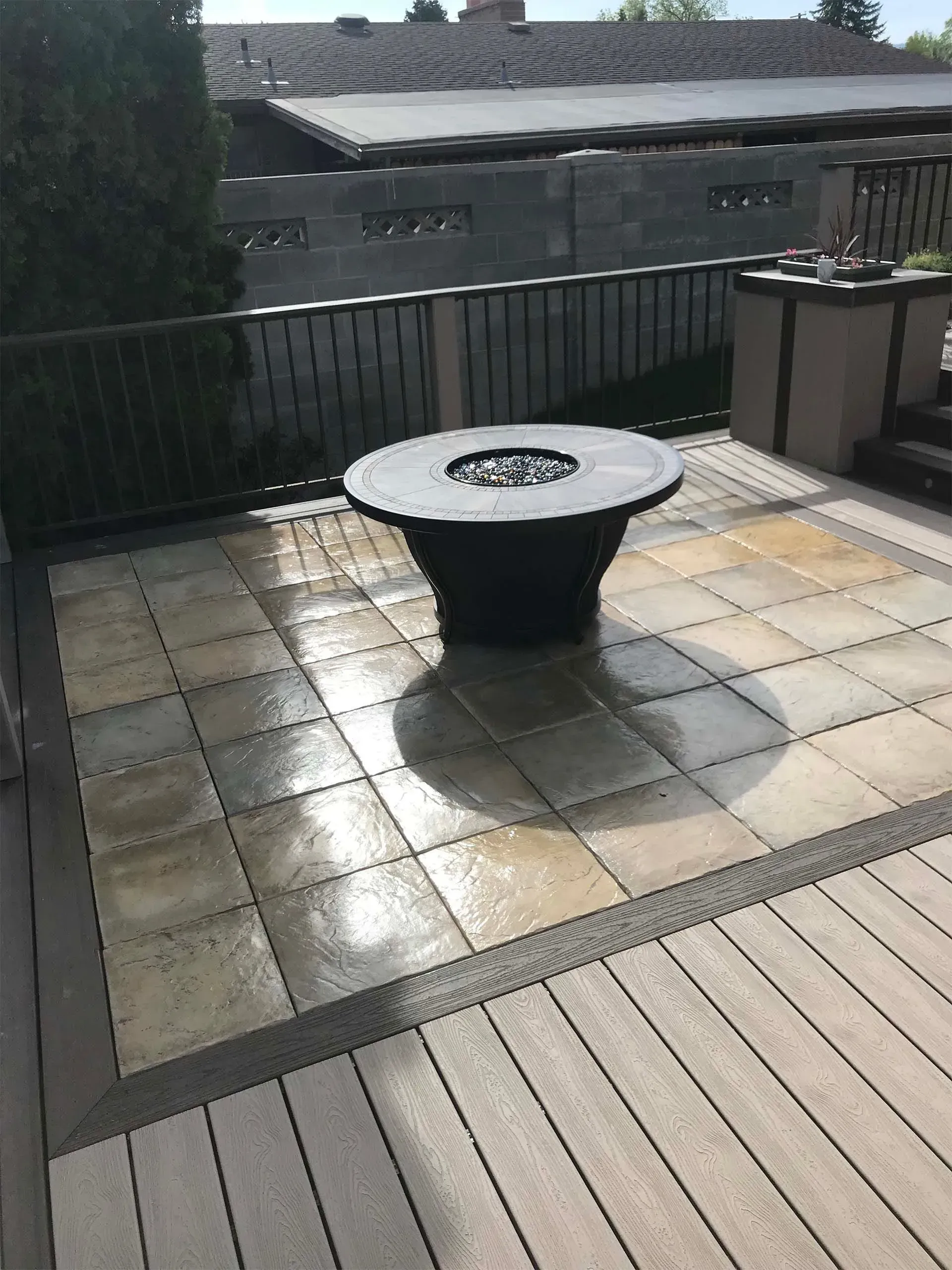 Patio with a dark fire pit on gray and tan stone pavers, surrounded by composite decking and a railing.