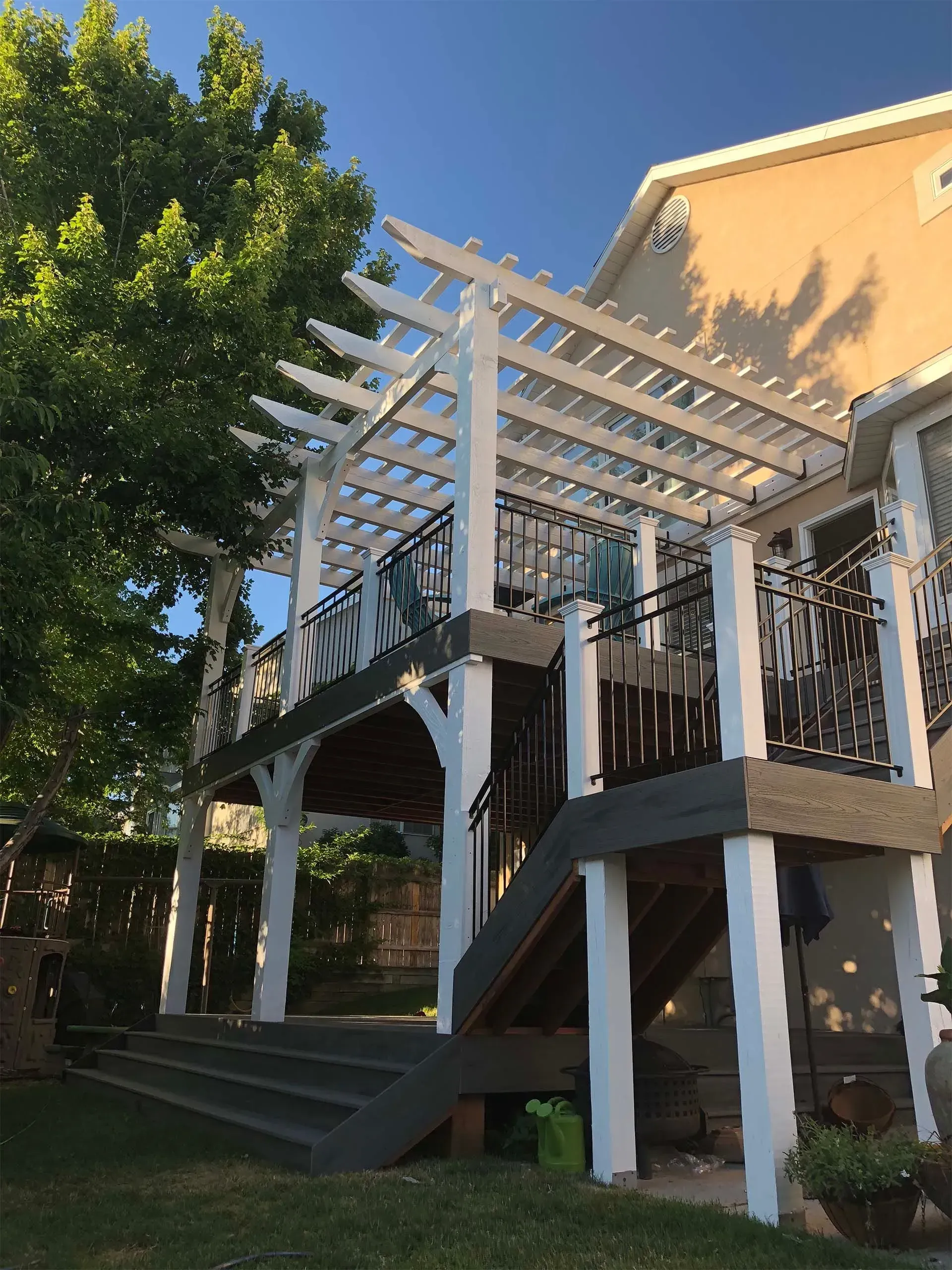 White pergola on a raised deck with steps. Black railing and tree in the background, sunny day.