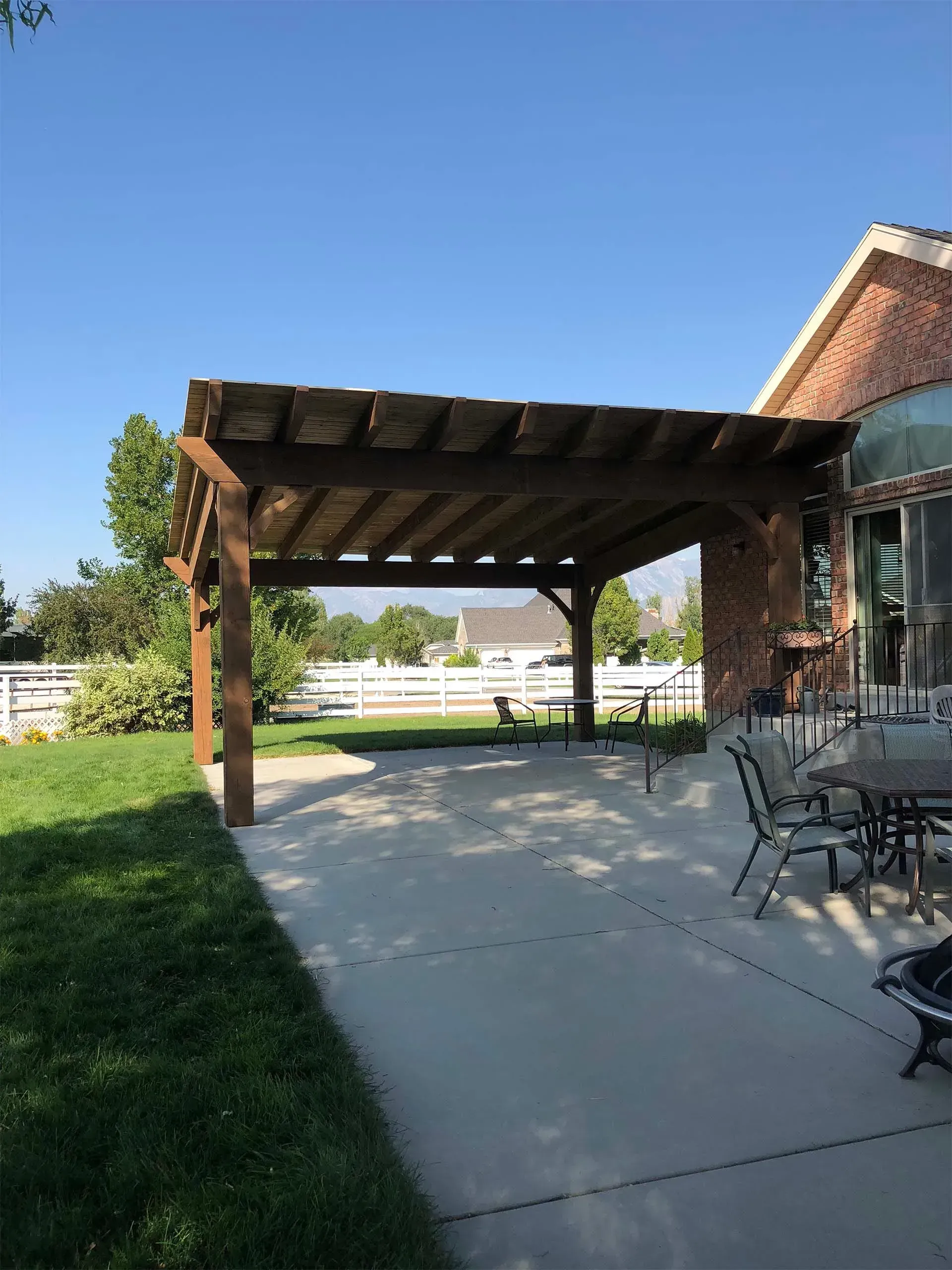 Wooden pergola over a concrete patio next to a brick house and green lawn on a sunny day.