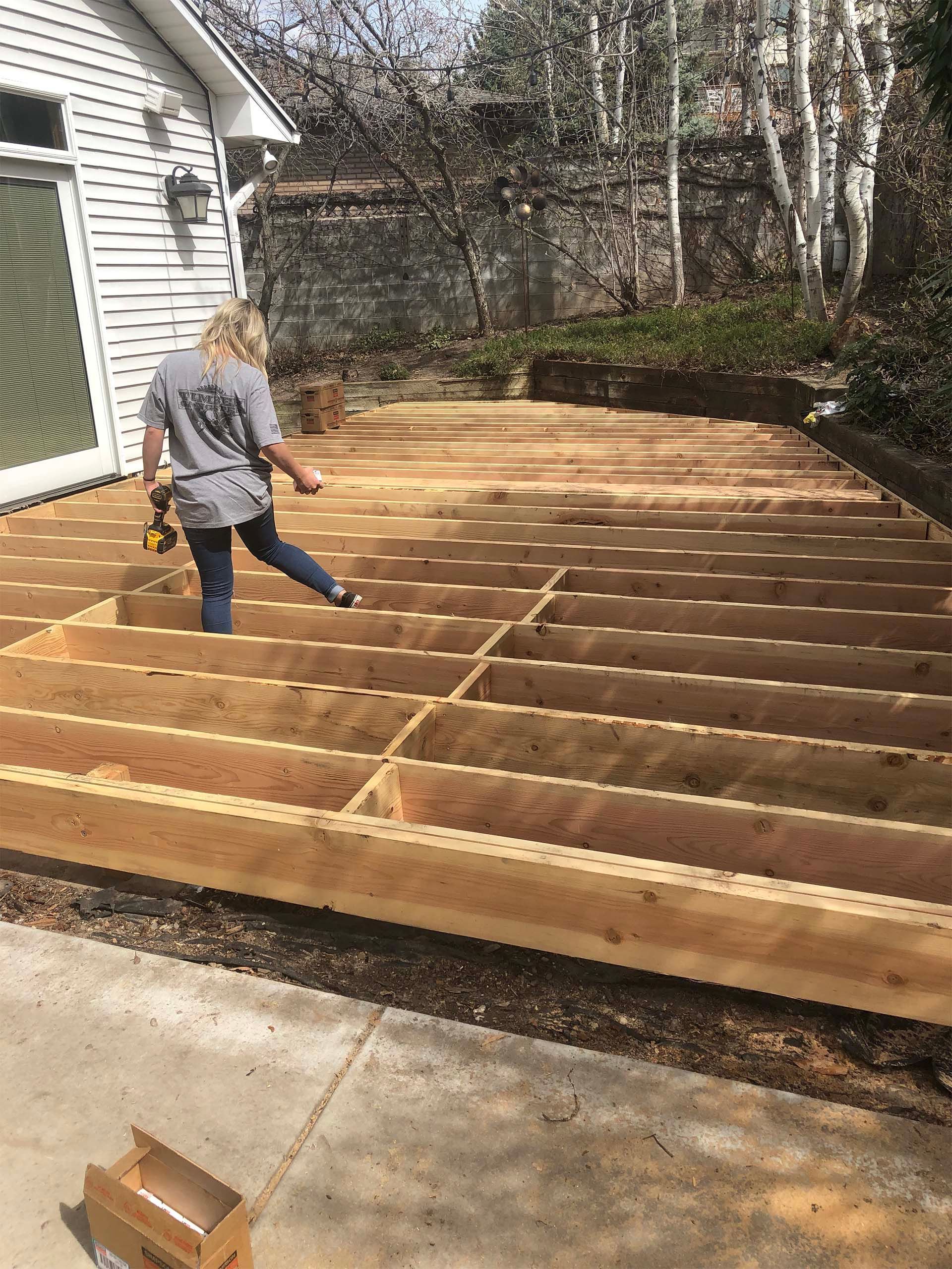 Woman building a deck outdoors; blond hair, holding drill, walking on exposed wooden frame.