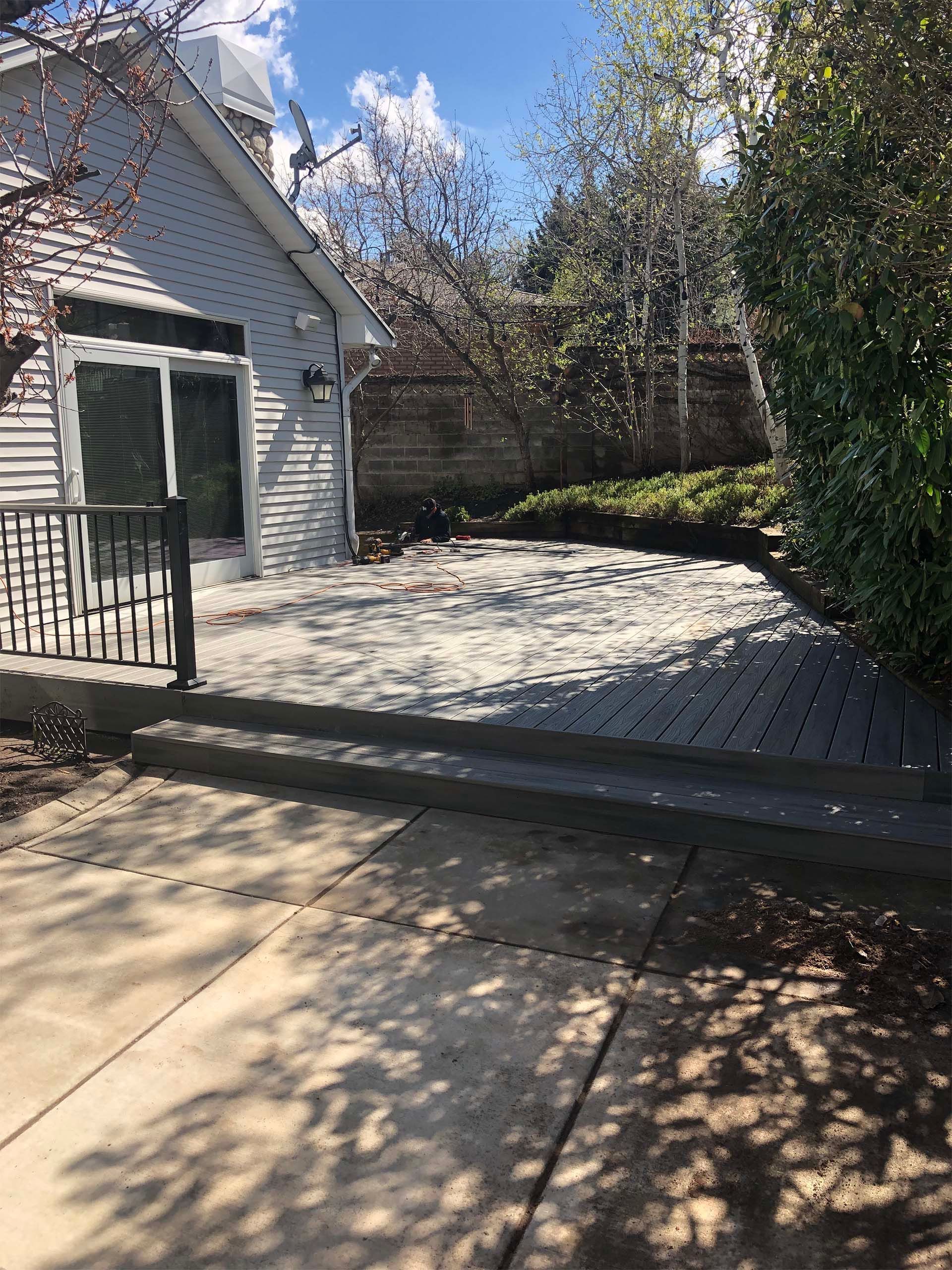 Backyard deck and patio, light gray and concrete with greenery and building.
