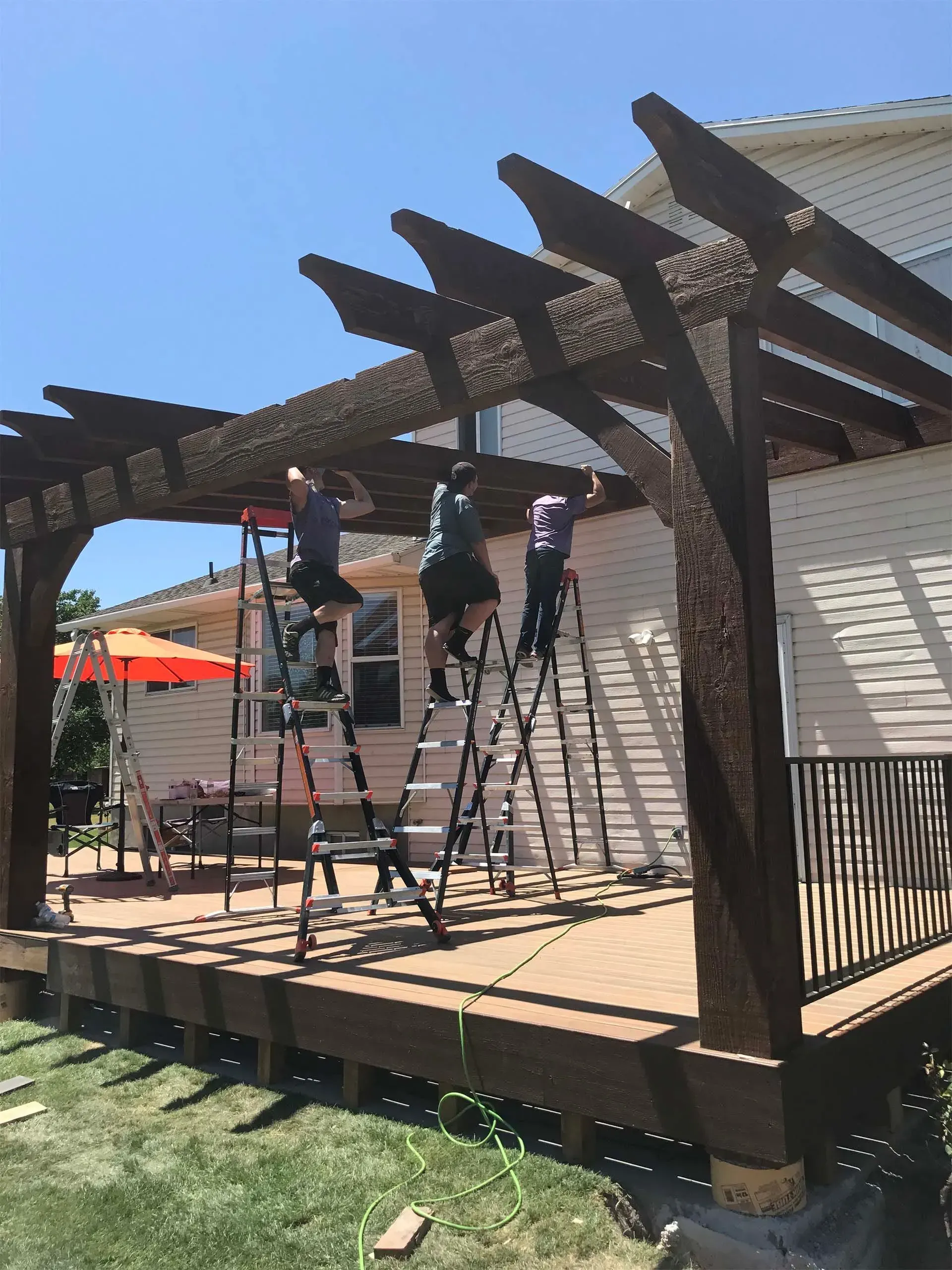 Three people working on a brown pergola, on a deck beside a house, using ladders.