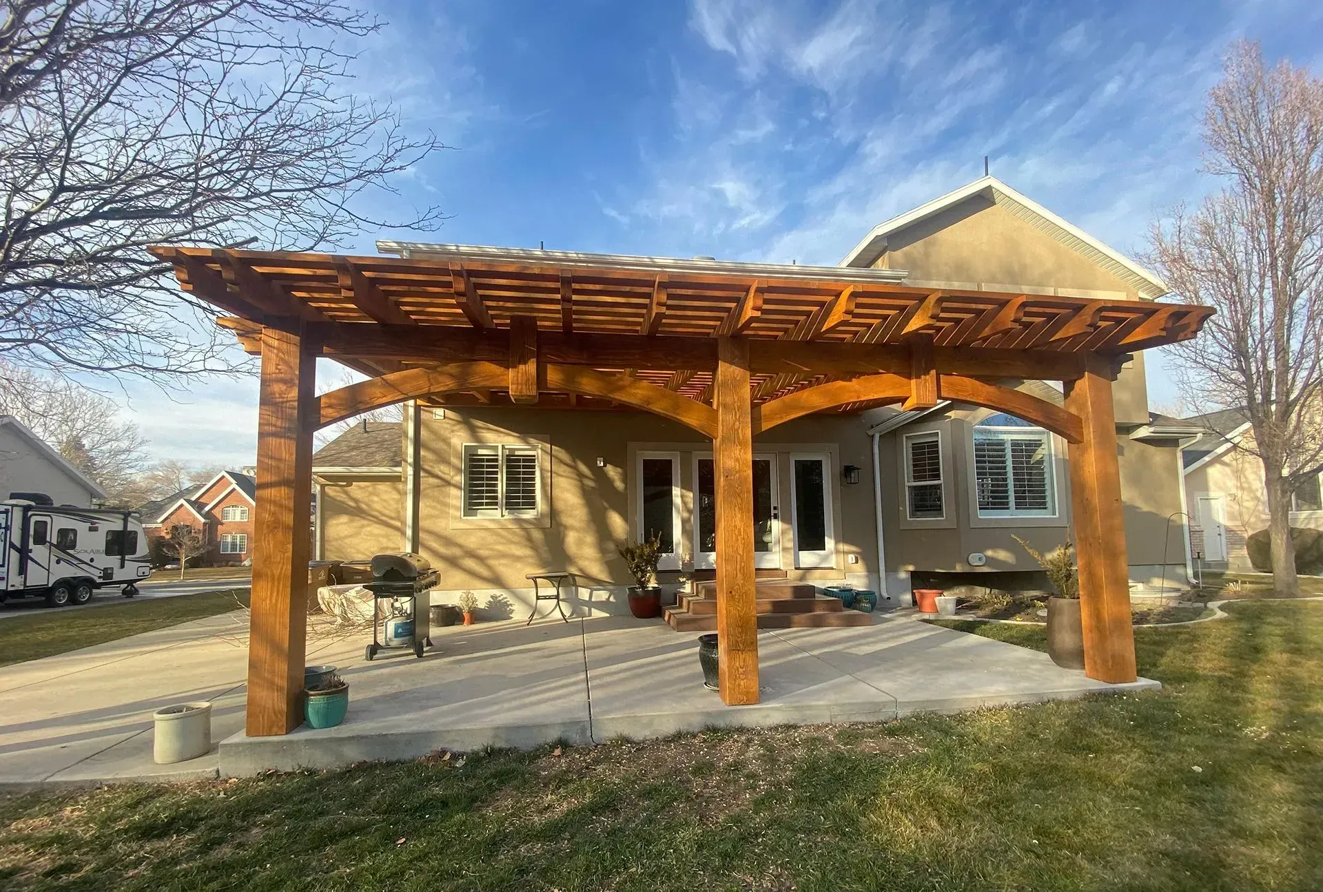 Wooden pergola over a concrete patio, attached to a tan house. Blue sky.