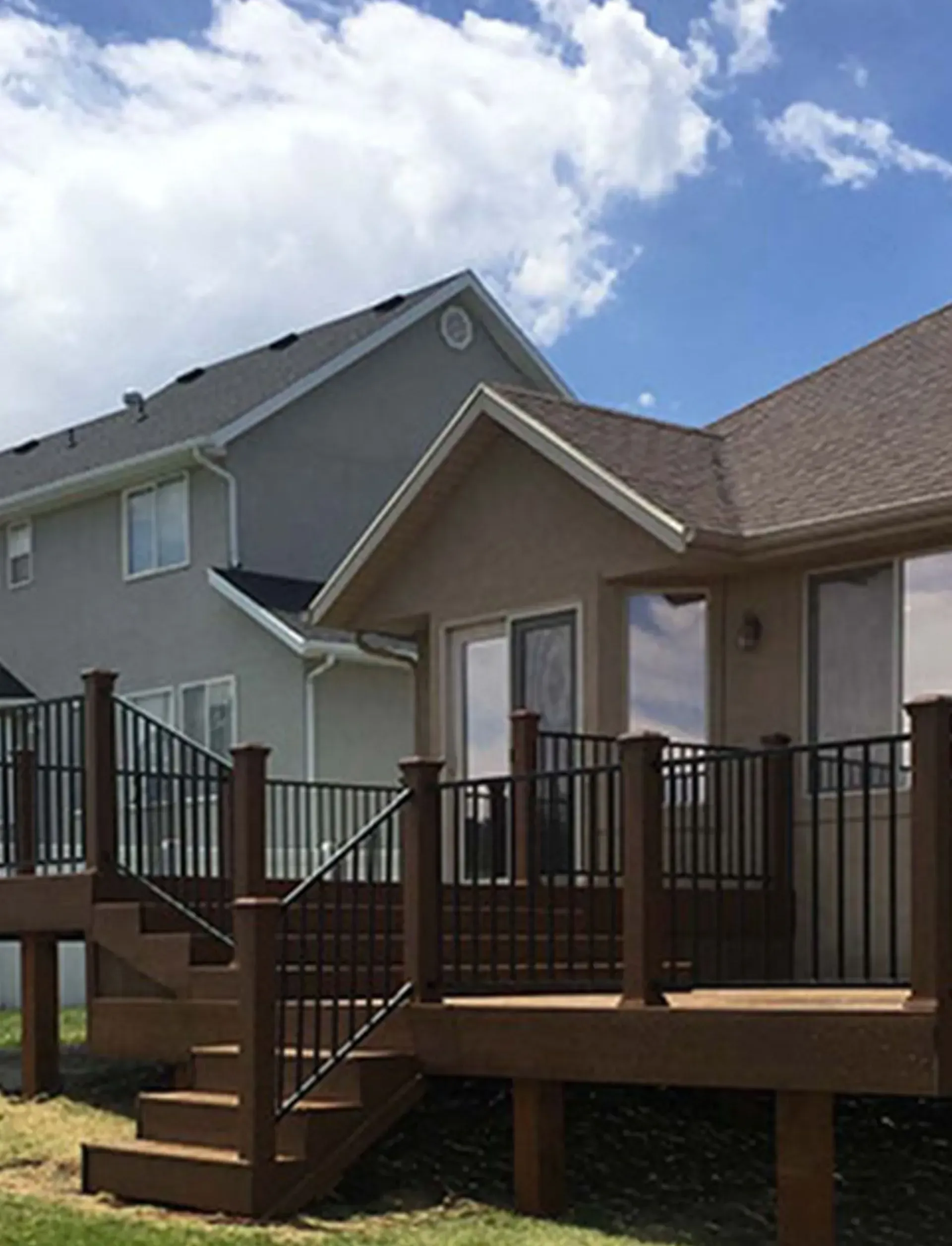 Deck with dark railings and steps in front of a two-story house with light gray siding and a blue sky.