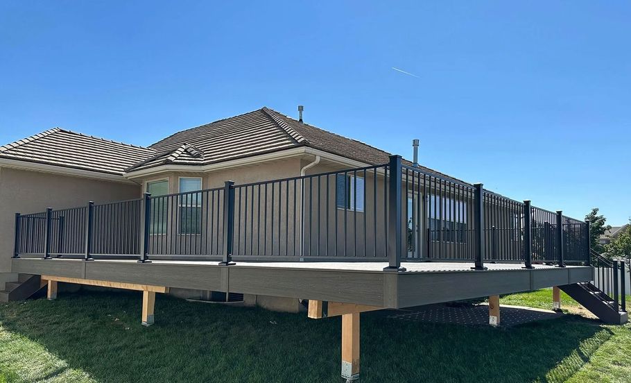 Elevated gray deck with black railing attached to a light-colored house on a sunny day.