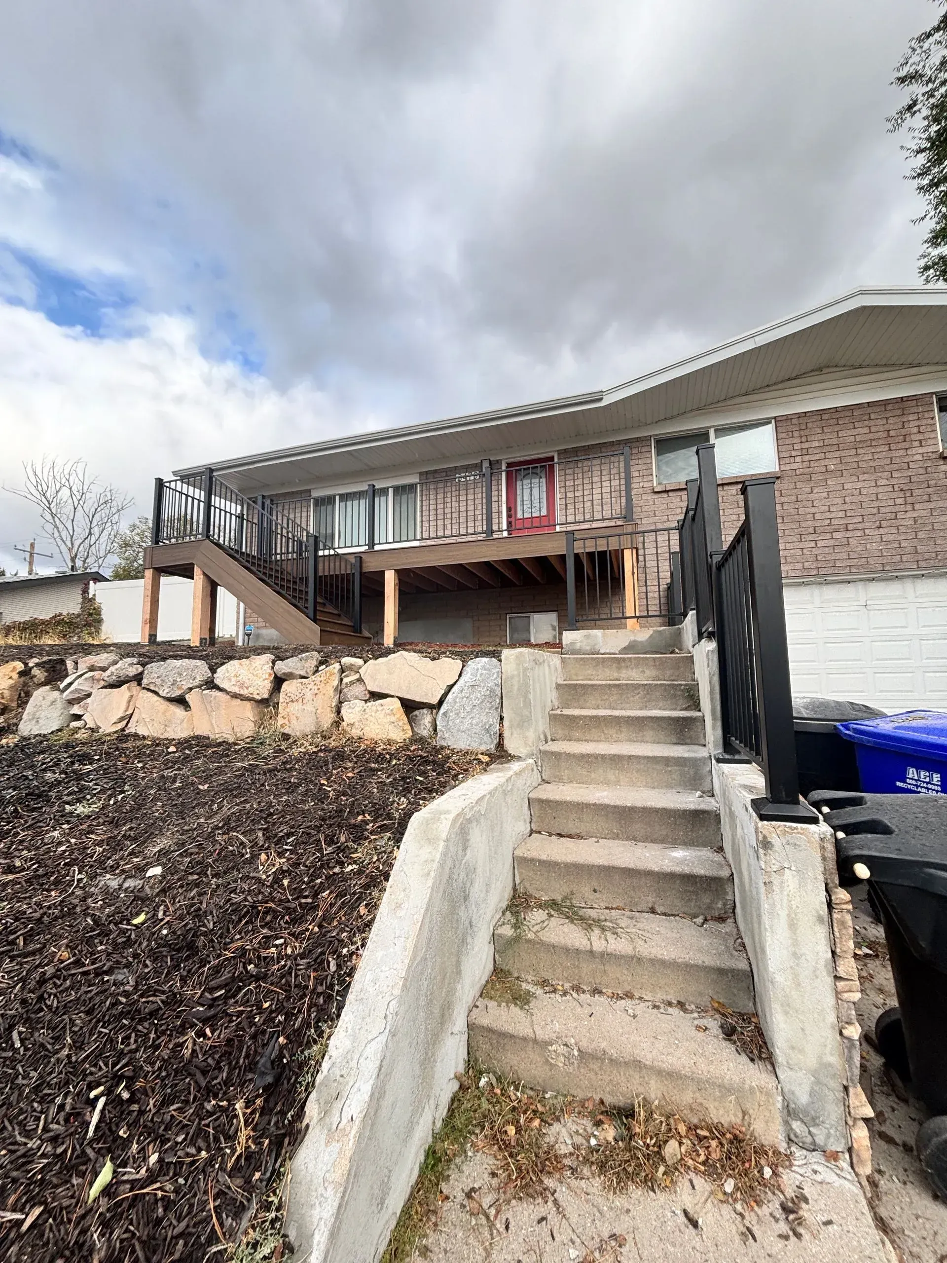 Concrete steps lead up to a house with a wooden deck and brown brick exterior on a cloudy day.