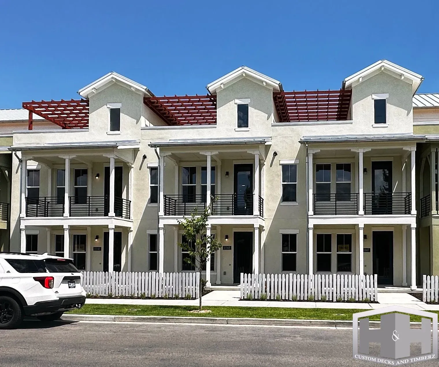 Row of townhouses with white picket fences, balconies, and a white SUV parked in front.