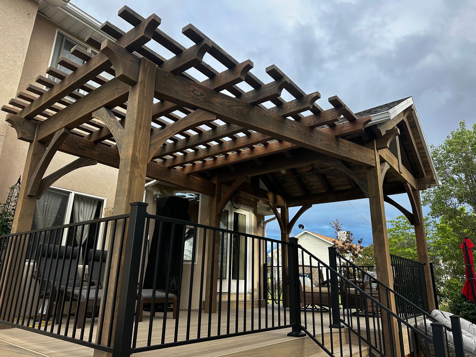 Wooden pergola over a patio with black railing and a cloudy sky.