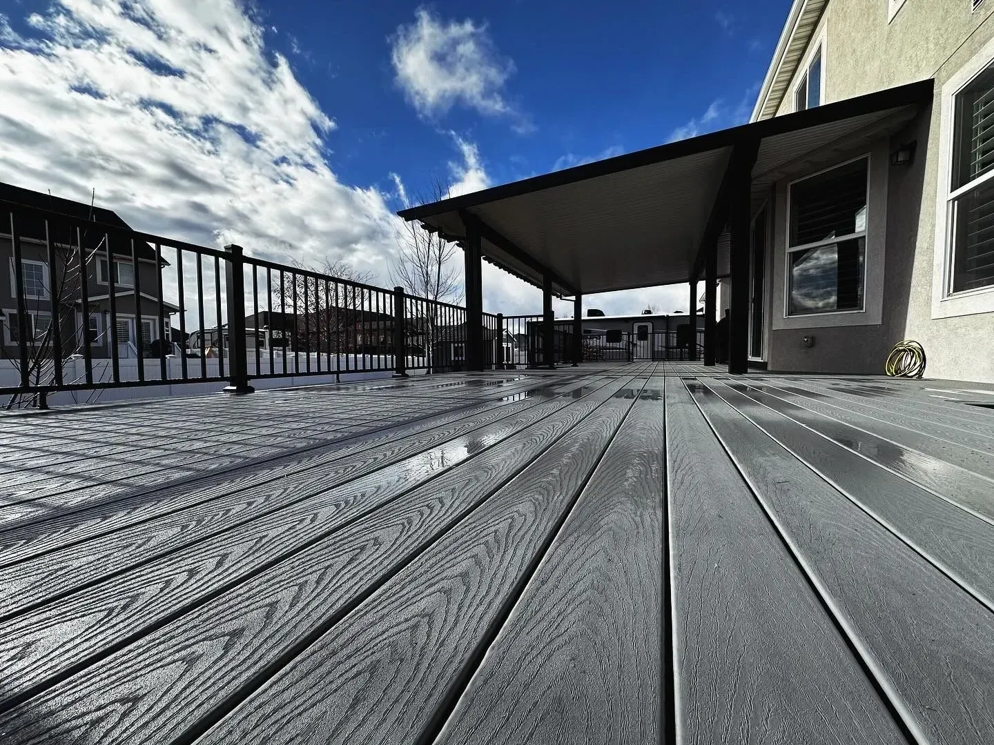 Gray composite deck extending from a beige house, under a covered patio. Bright blue sky with clouds.