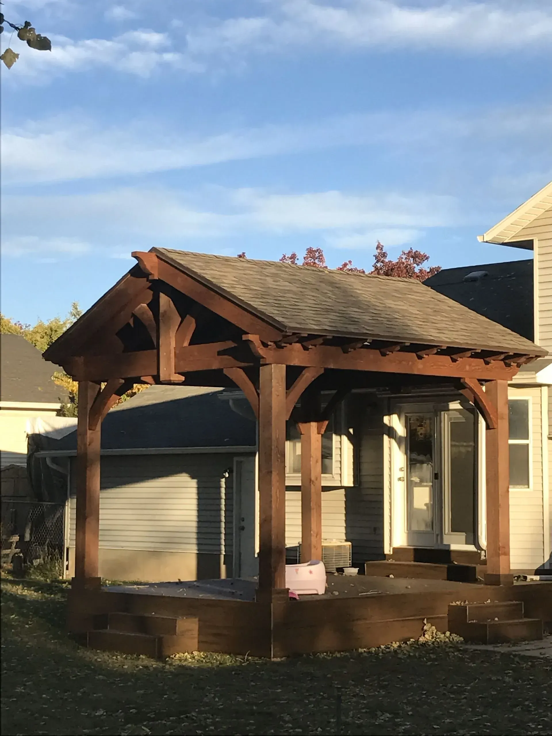 Wooden pergola with a shingled roof on a deck, near a house, under a blue sky.