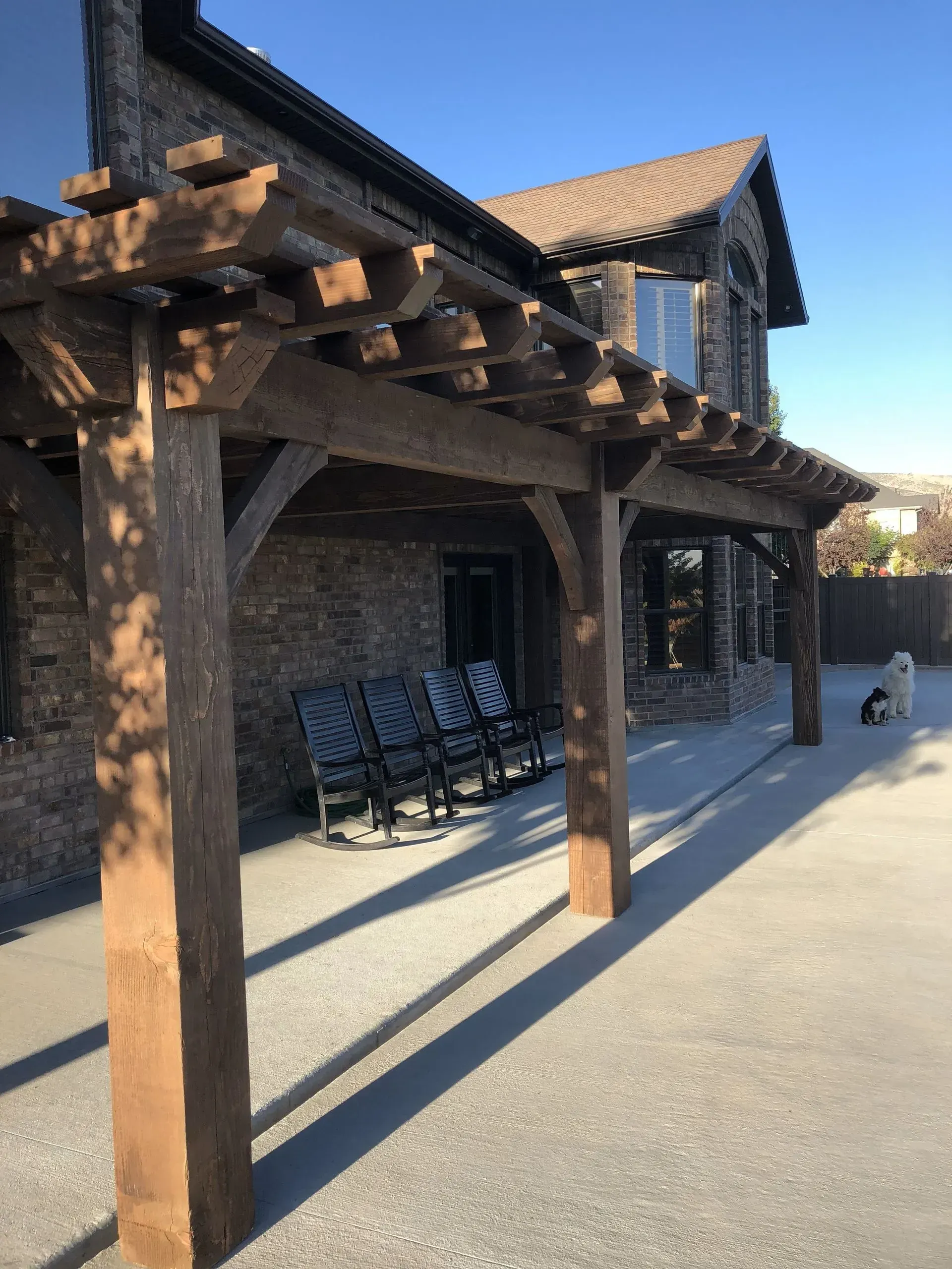 Wooden pergola shading a stone building's porch with rocking chairs. Sunny day, shadows cast.