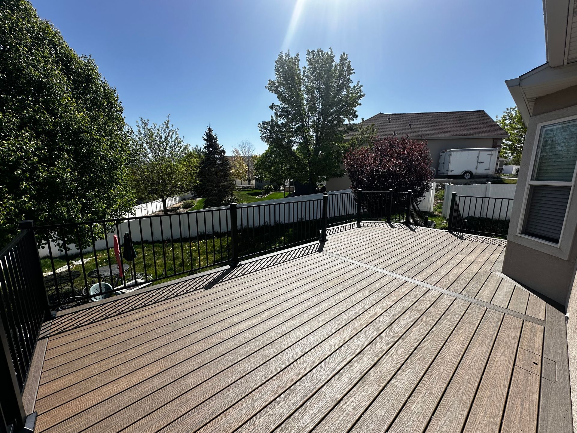 A wooden deck with black railings overlooks a backyard with trees, a fence, and blue sky.