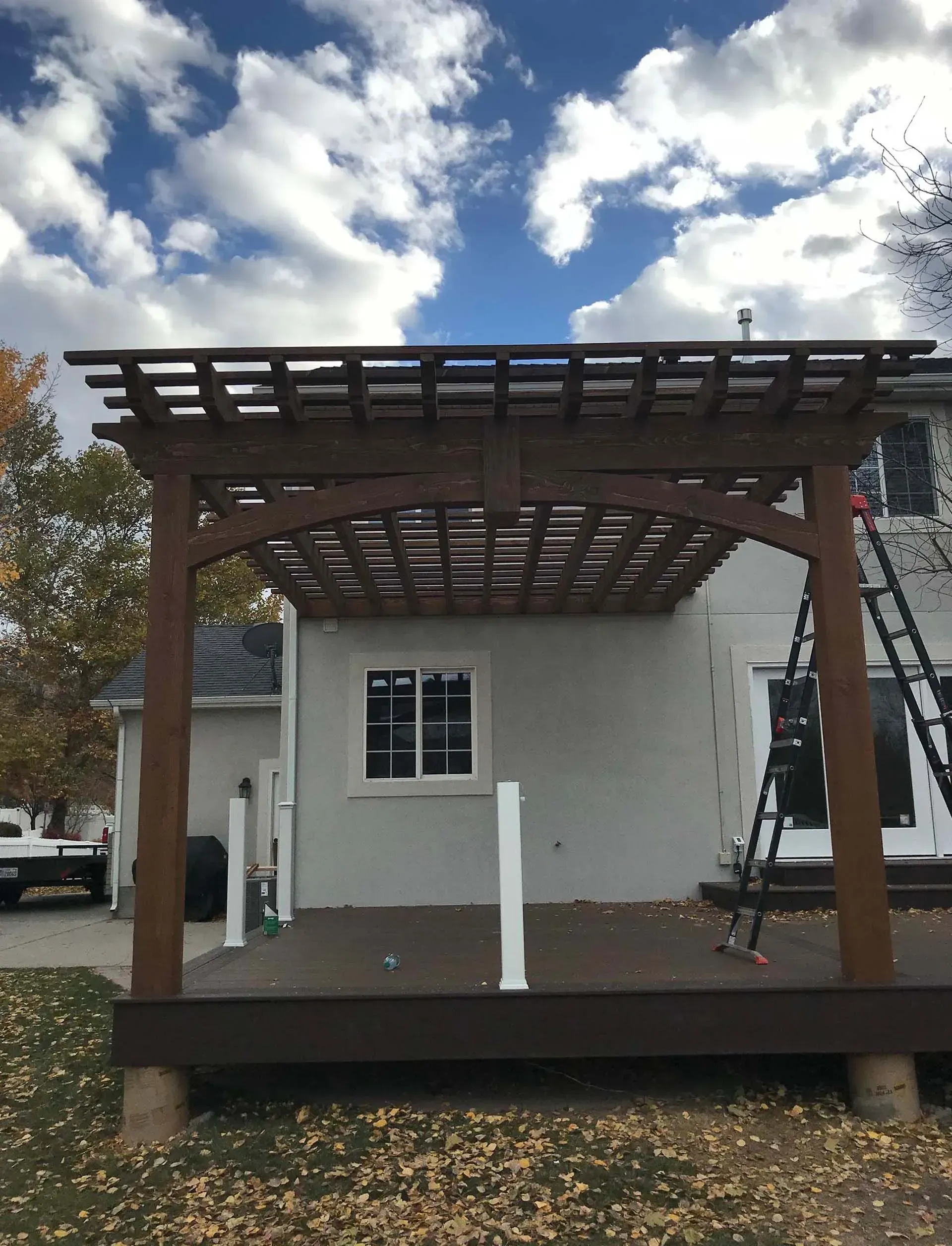 Wooden pergola over a deck on the back of a house. The sky is blue and cloudy.