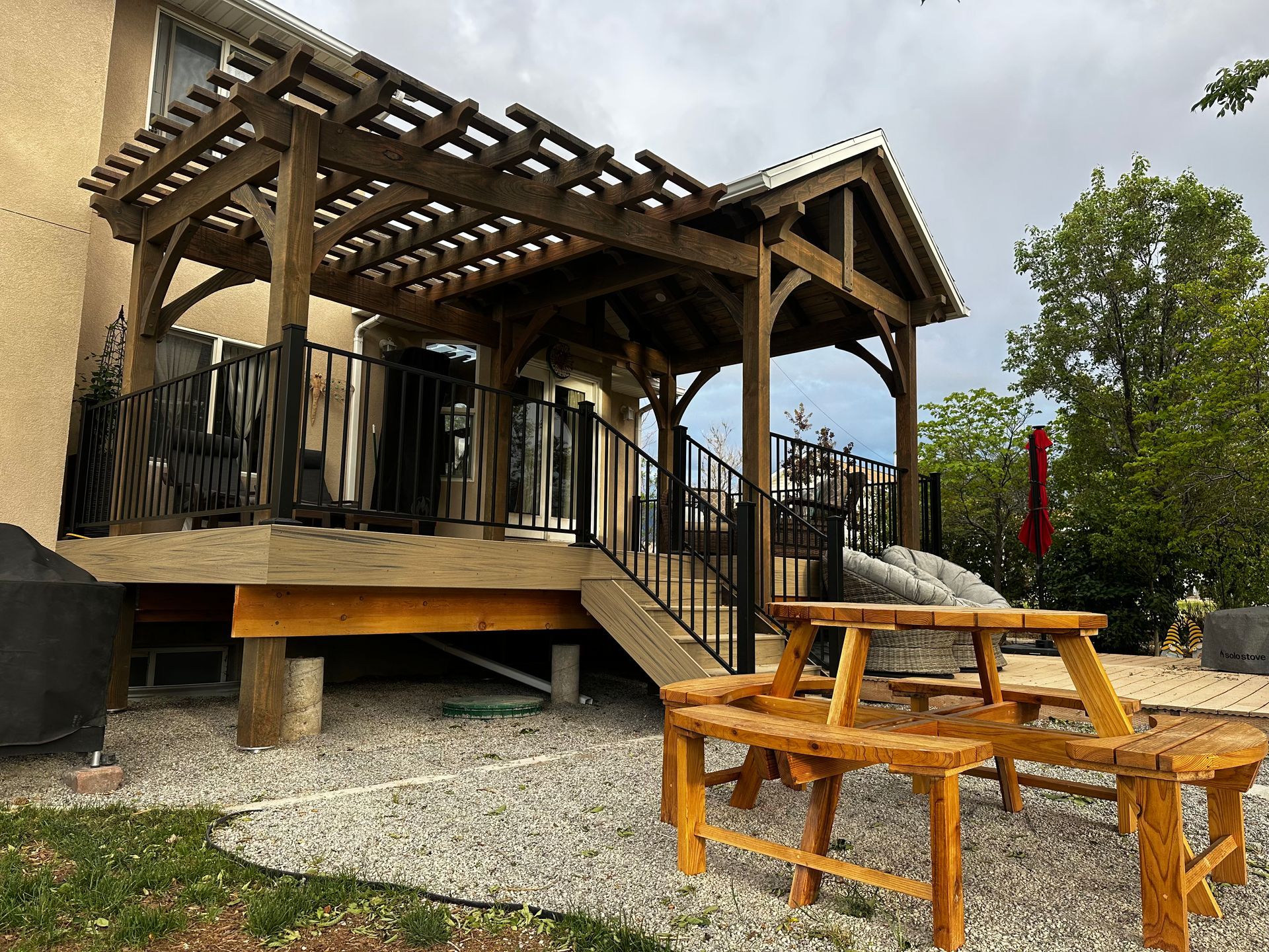 Wooden pergola on deck, with picnic table in a gravel yard.