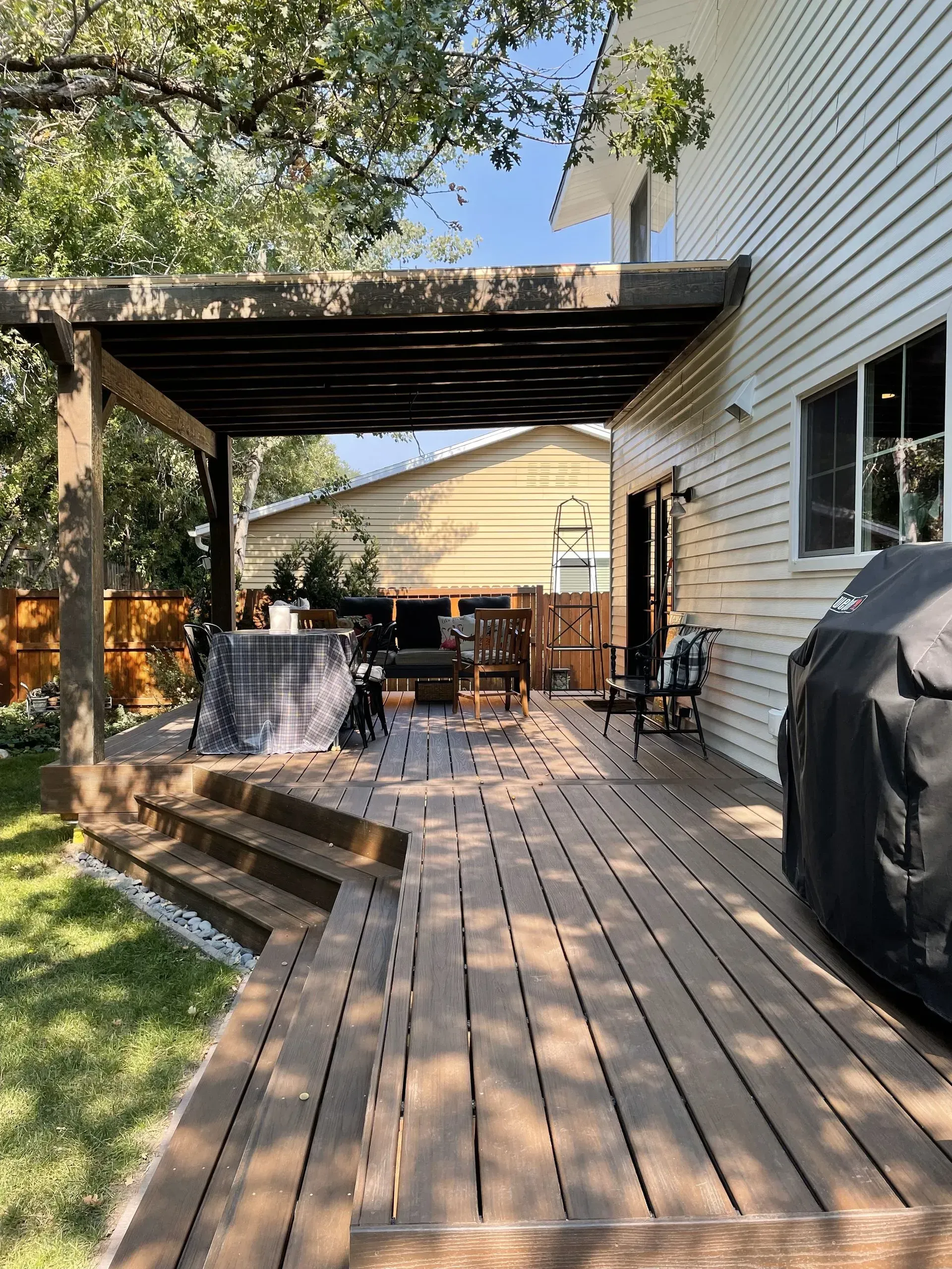 Wooden deck with pergola attached to a white house. Backyard with seating and a grill.