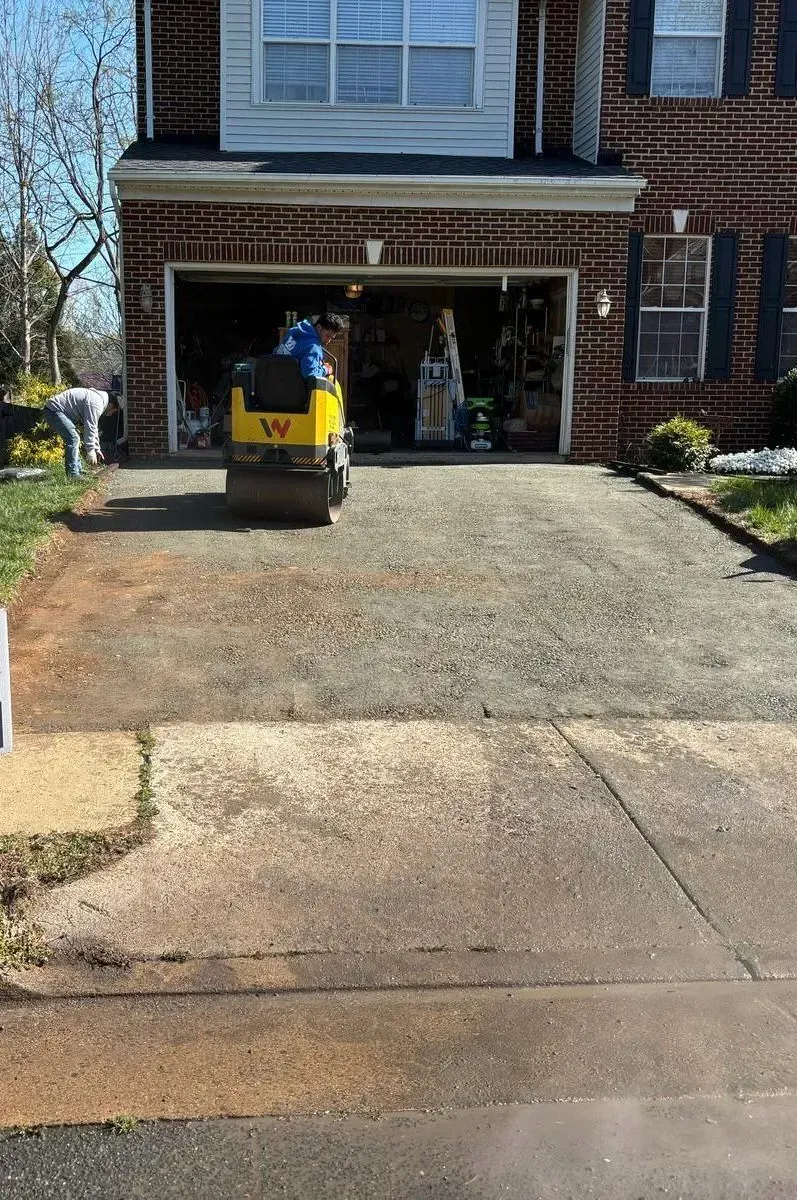 A roller is rolling concrete in a driveway in front of a brick house.