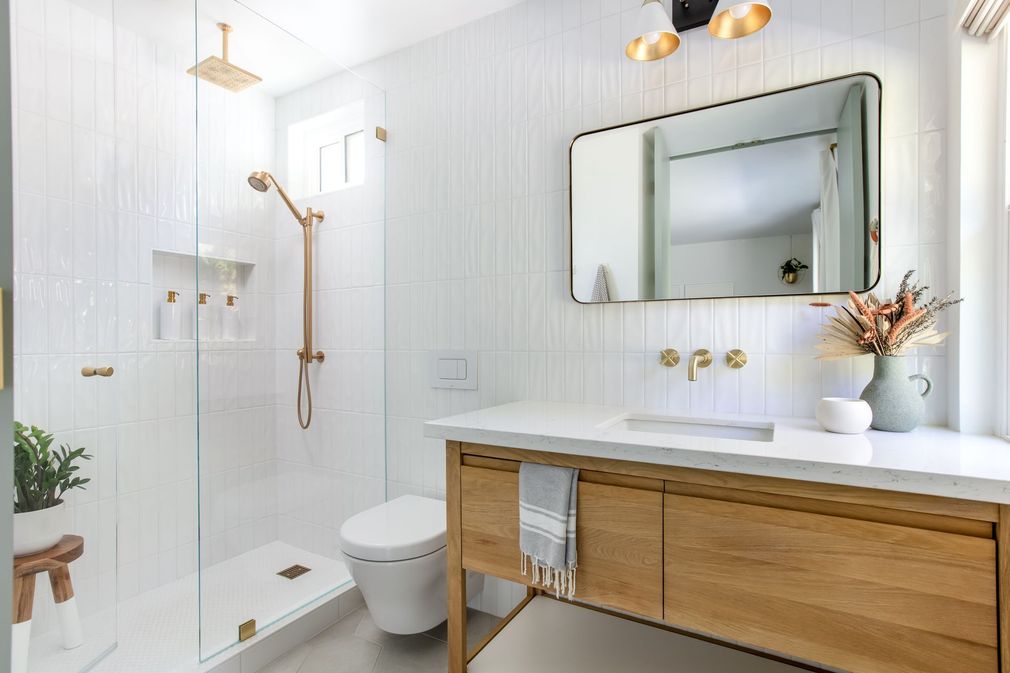 Bright white bathroom with glass shower, wooden vanity, round mirror, and brass fixtures