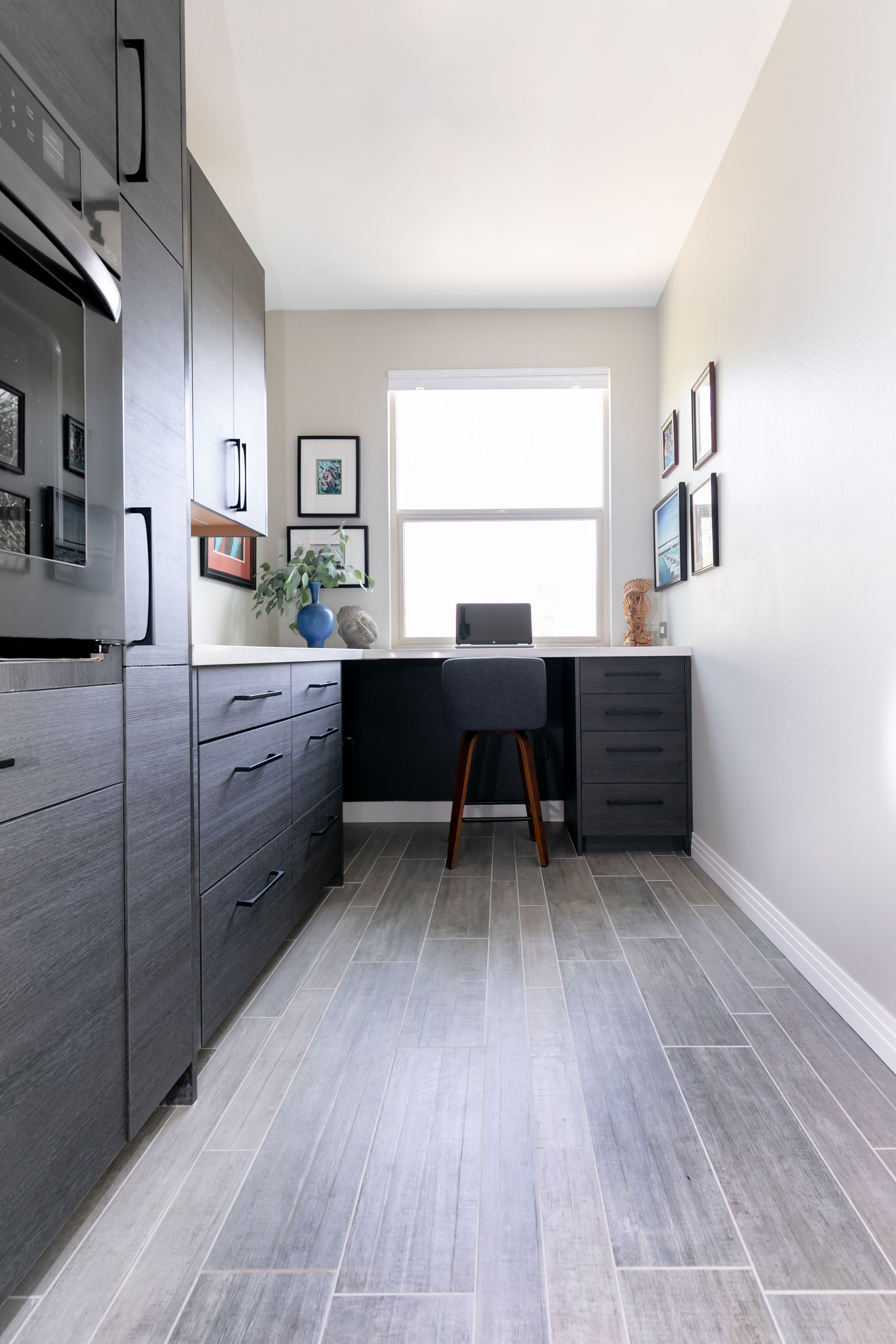 Narrow home office with dark cabinets, desk by a bright window, and light gray tile floor.