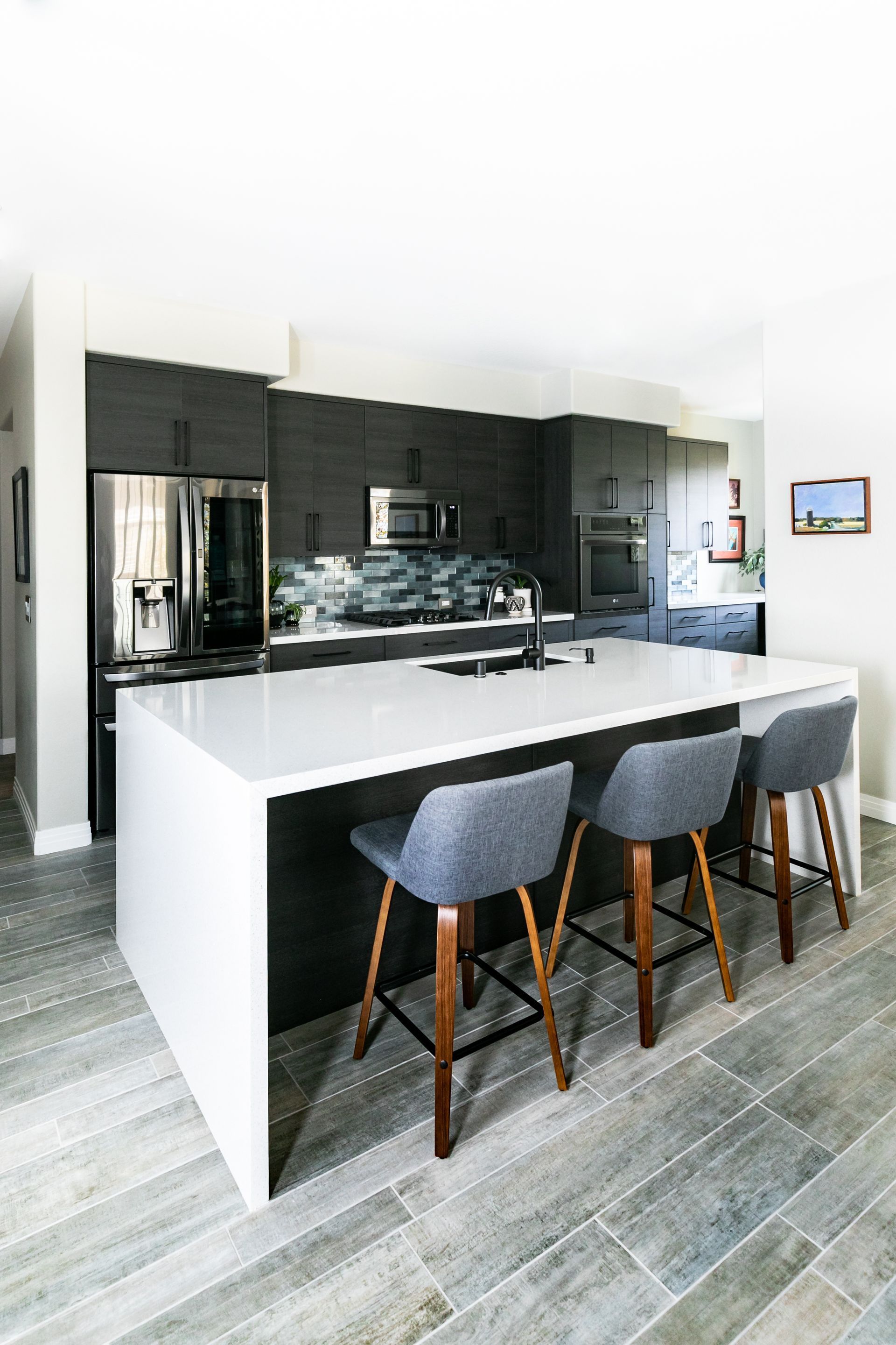 Modern kitchen with white island, dark cabinets, and three gray bar stools on gray wood flooring