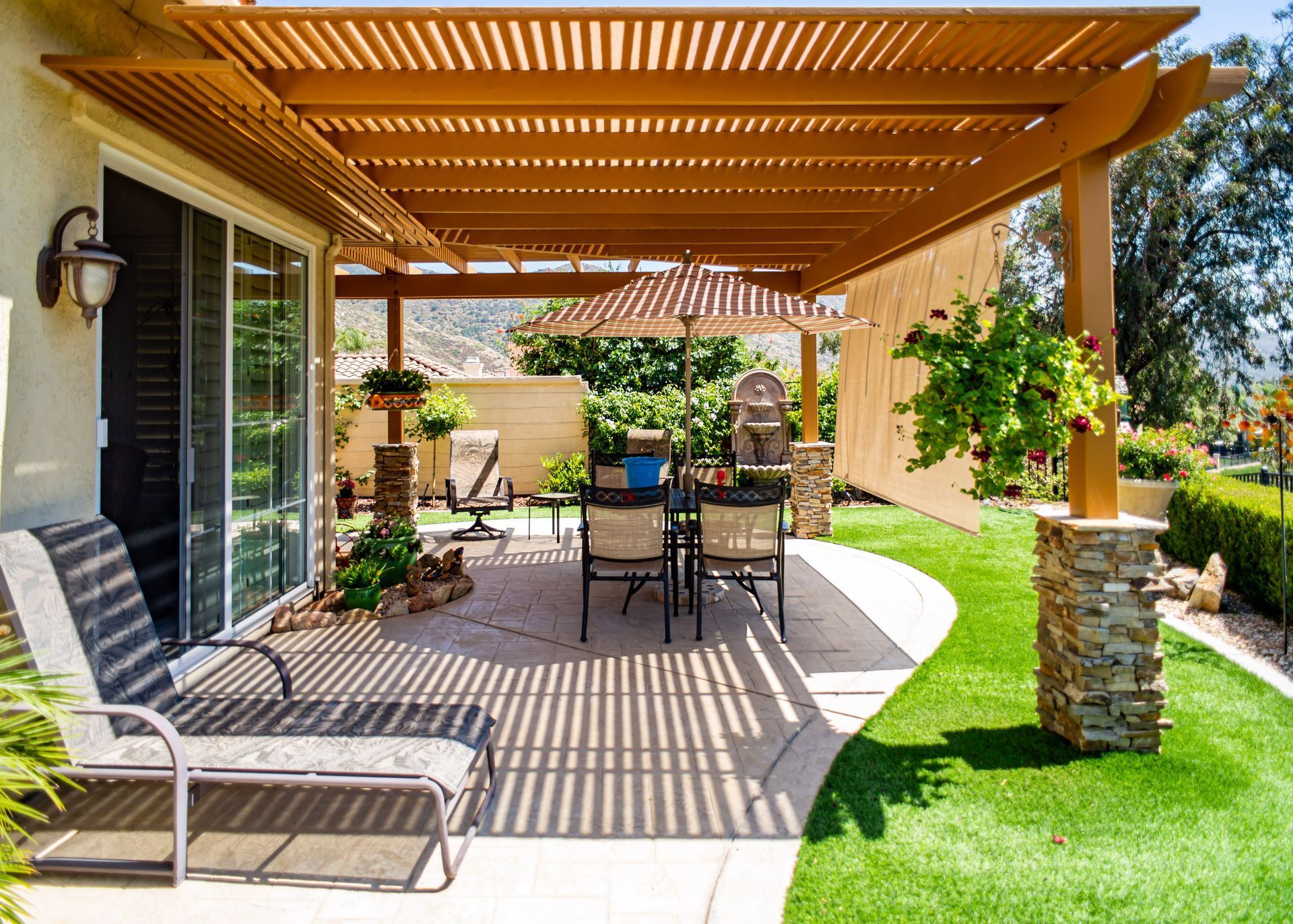 Covered patio with wood pergola, outdoor dining table, lounge chair, and potted plants on a sunny lawn
