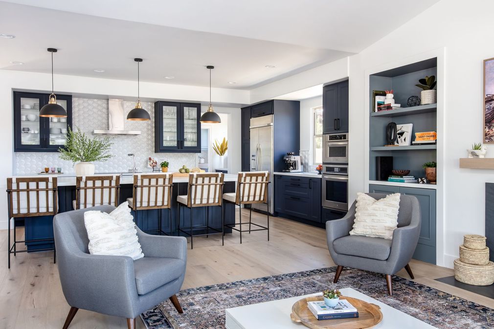 Bright modern living room with gray chairs, rug, and open kitchen with dining table and dark cabinets.