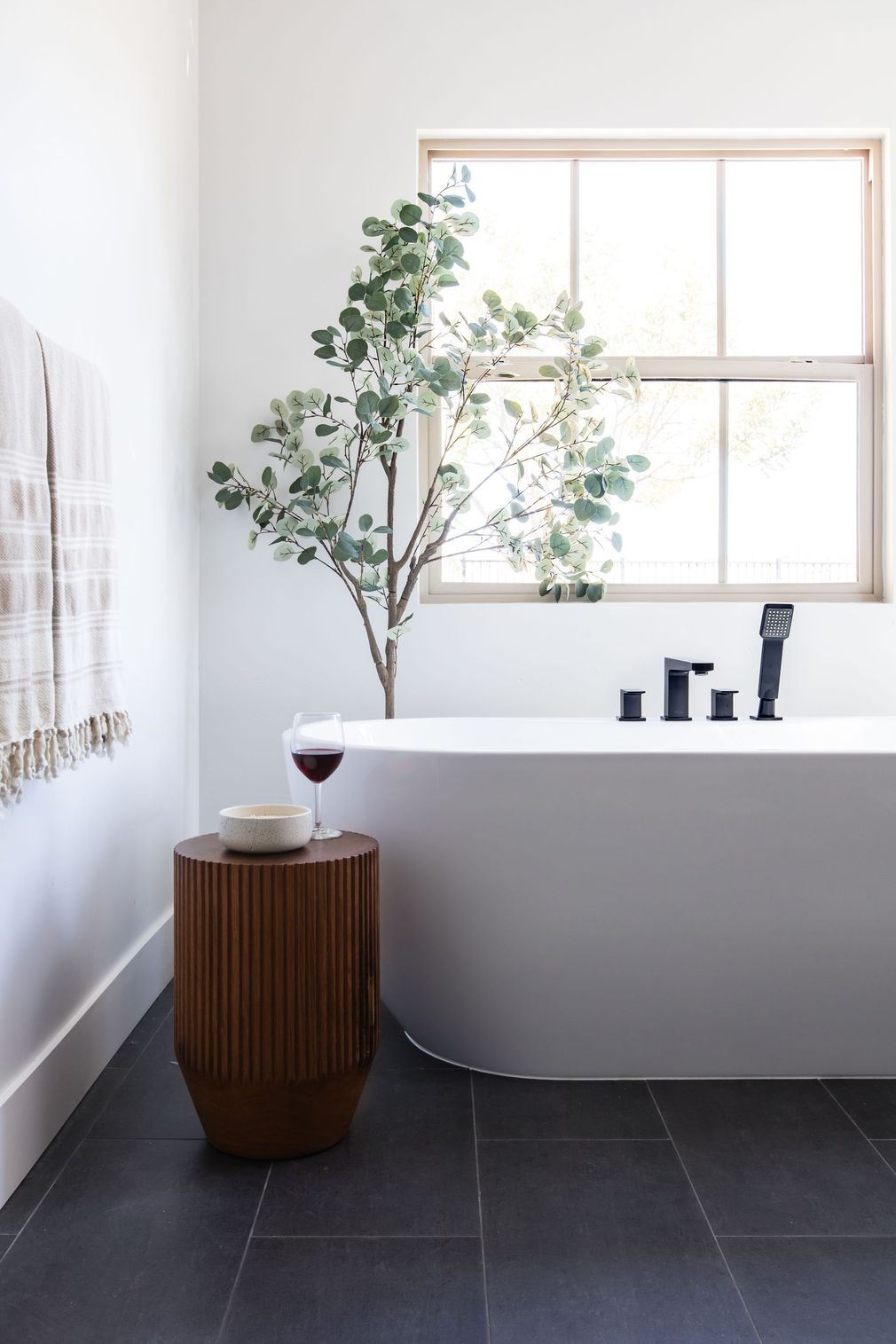 Modern bathroom with freestanding tub, dark tile floor, wood stool, and potted tree by a window