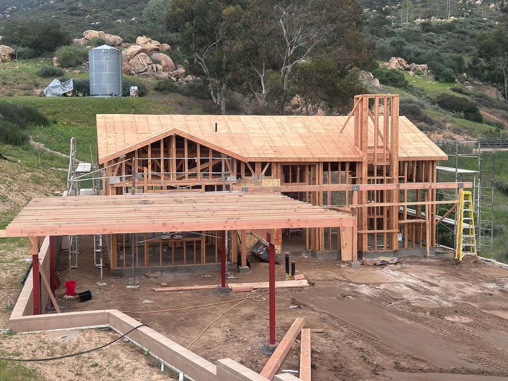 Partially built wooden house with scaffolding on a dirt lot, surrounded by trees and hills
