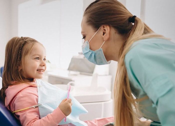 A little girl is sitting in a dental chair with a dentist wearing a mask.