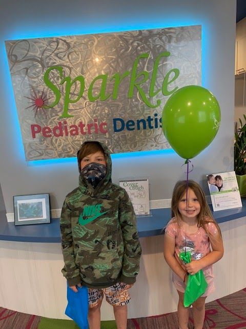 A boy and a girl are standing in front of a sparkle pediatric dentist sign.