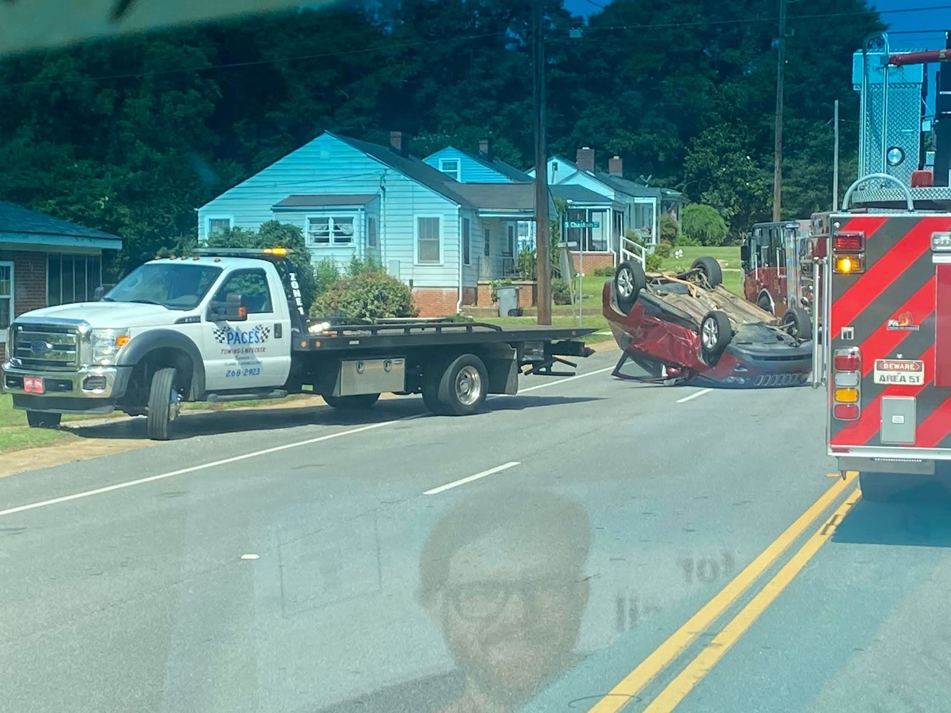 Tow truck at scene of car accident; overturned red car. Fire truck, houses and trees visible.