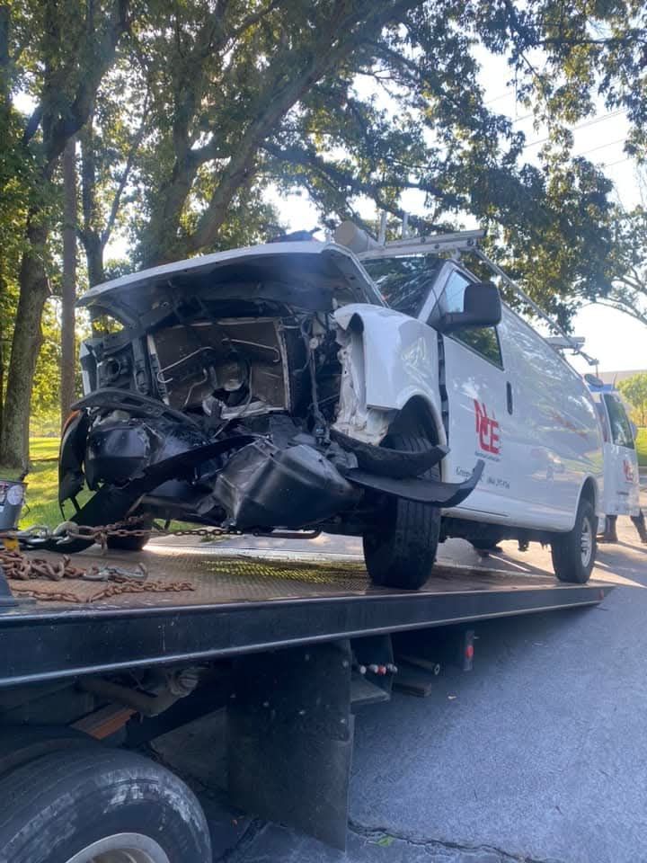 Damaged white van on a tow truck, front end heavily crushed in a crash.