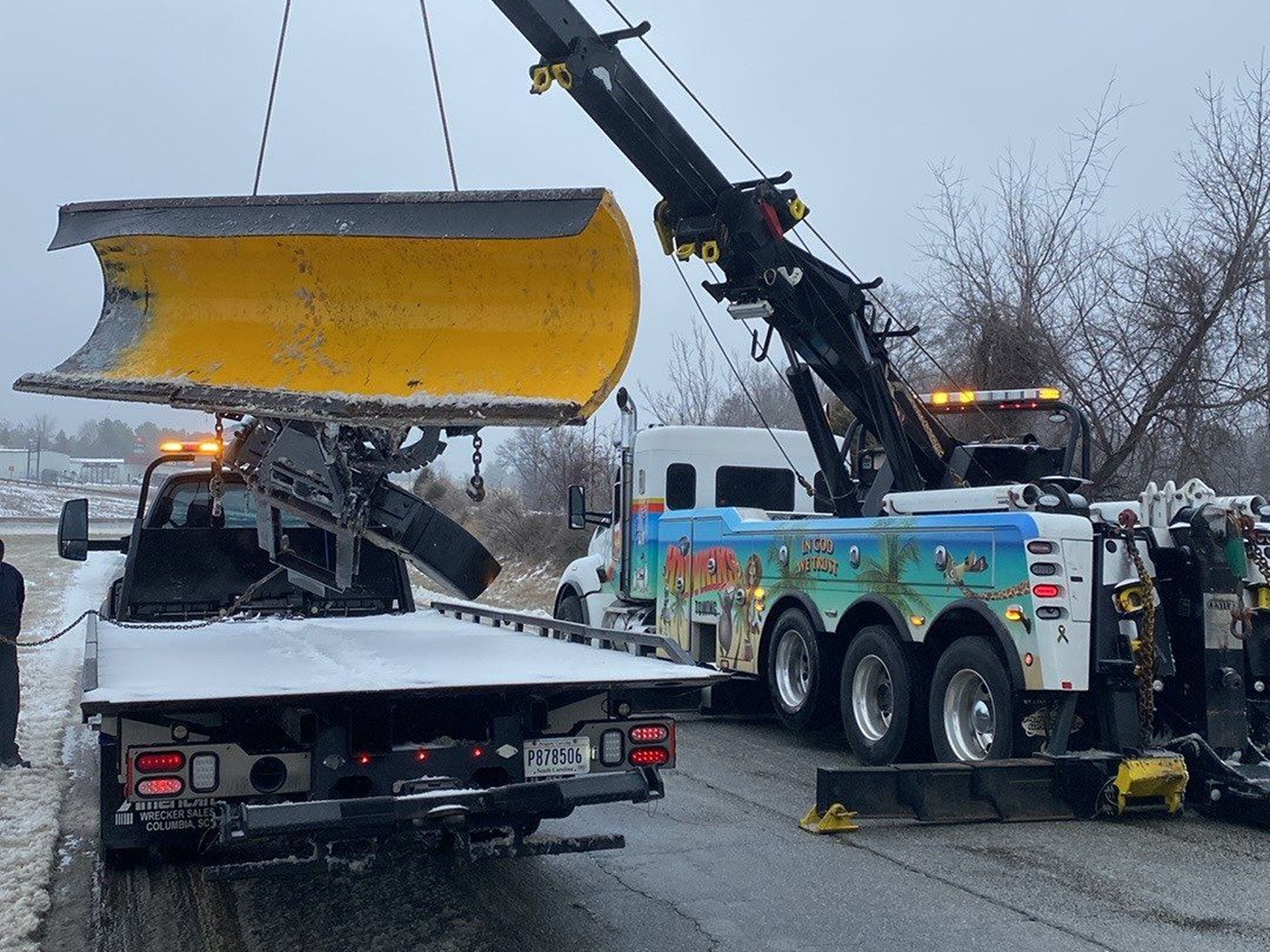 Tow truck lifting a yellow snowplow blade in a snowy roadside scene
