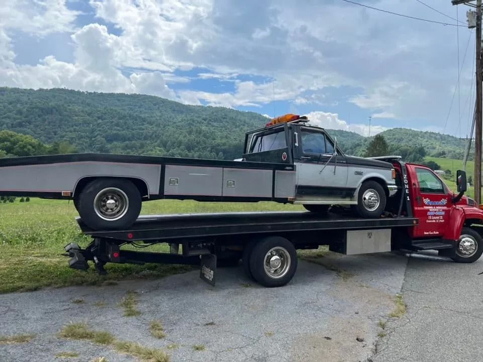 Tow truck transporting a silver and black pickup truck on a roadside, mountains in the background.