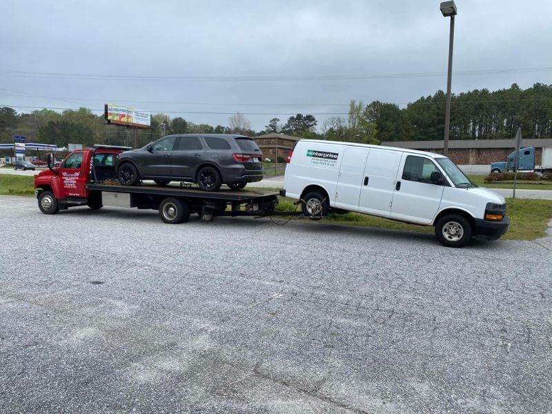Tow truck carrying a gray SUV and white van on a flatbed, parked on gravel.