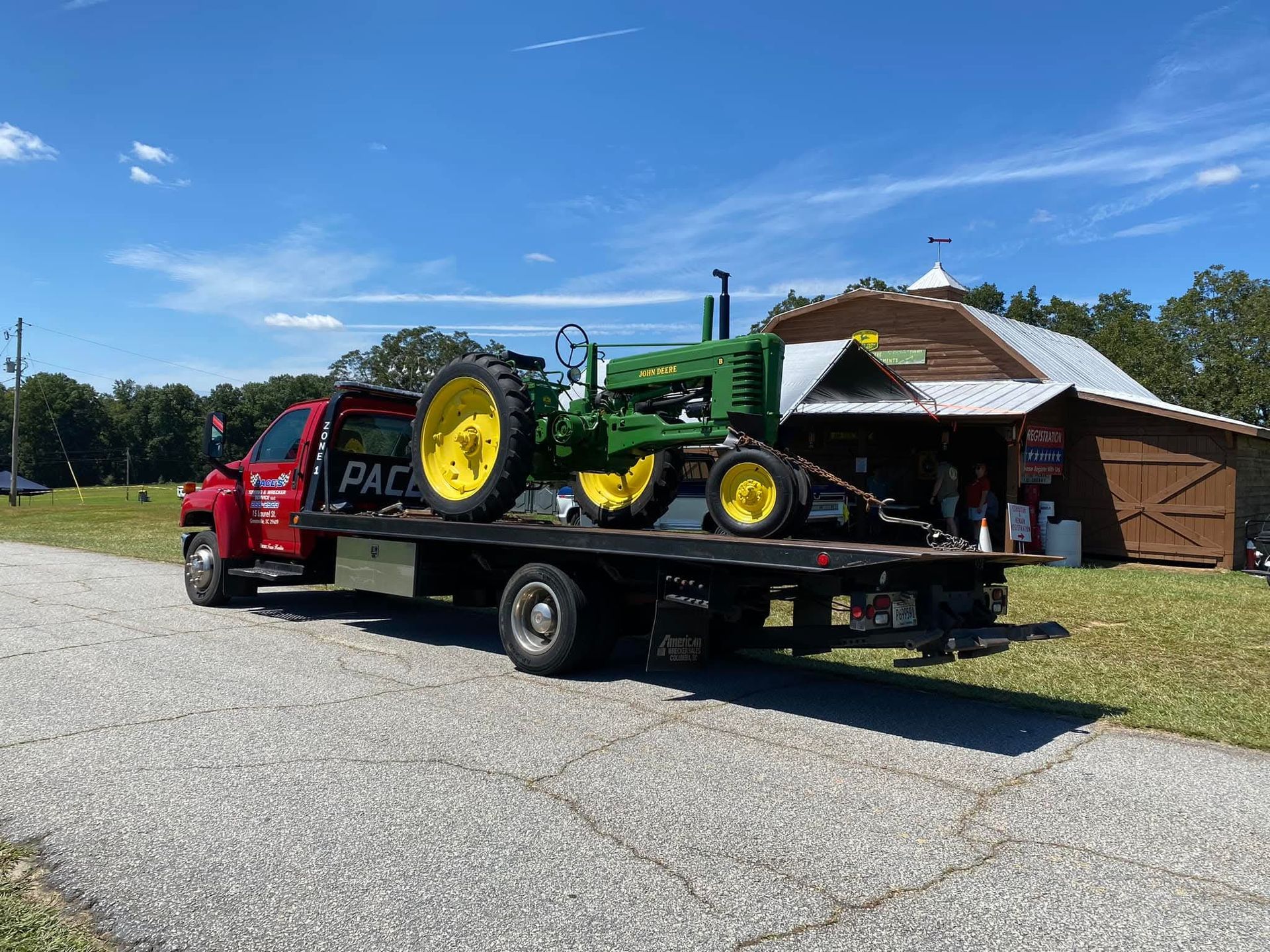 A green and yellow John Deere tractor on a flatbed tow truck in front of a barn.