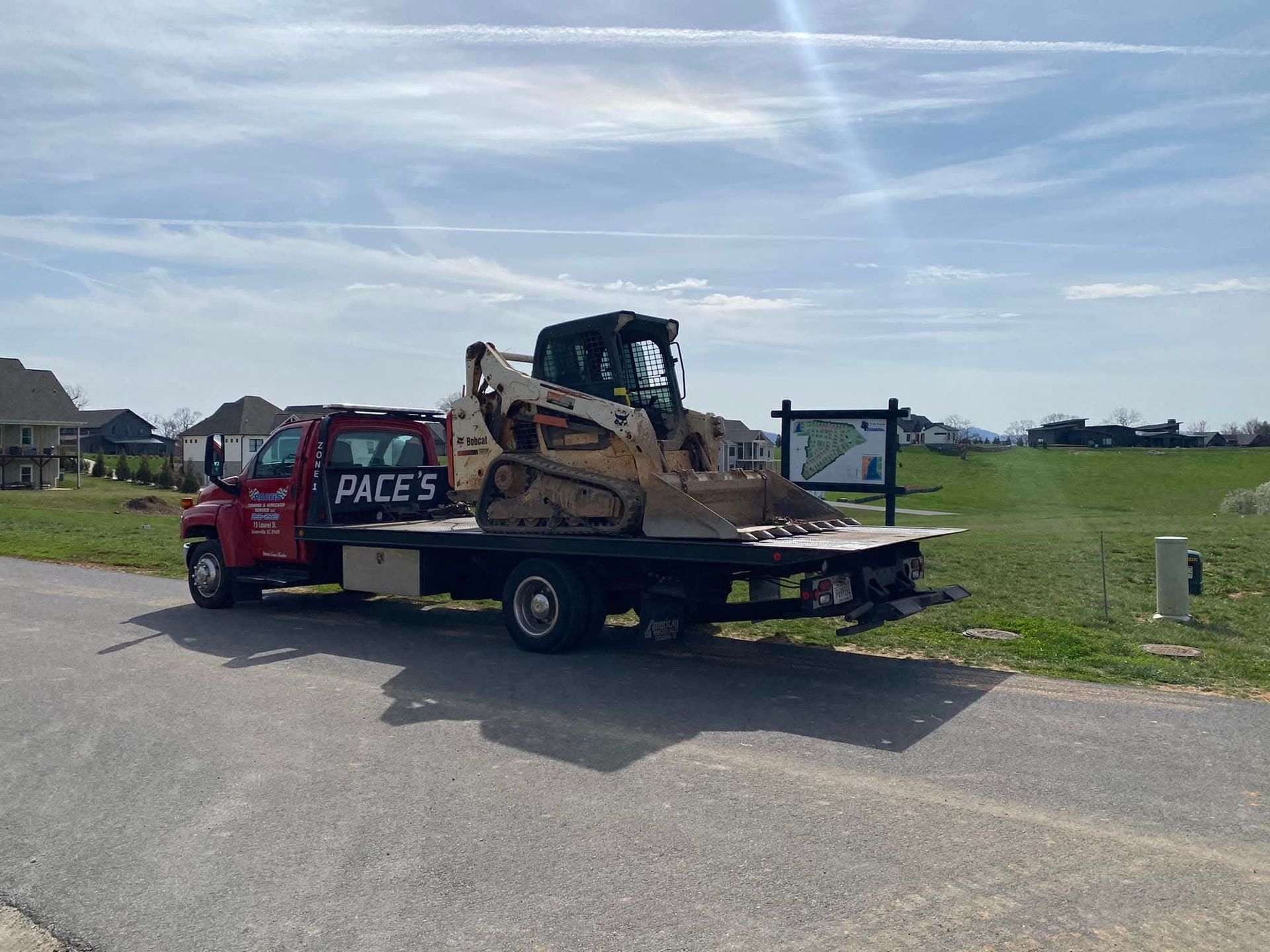 Red Pace's truck carrying a skid steer loader on a paved road, houses and a field in background.