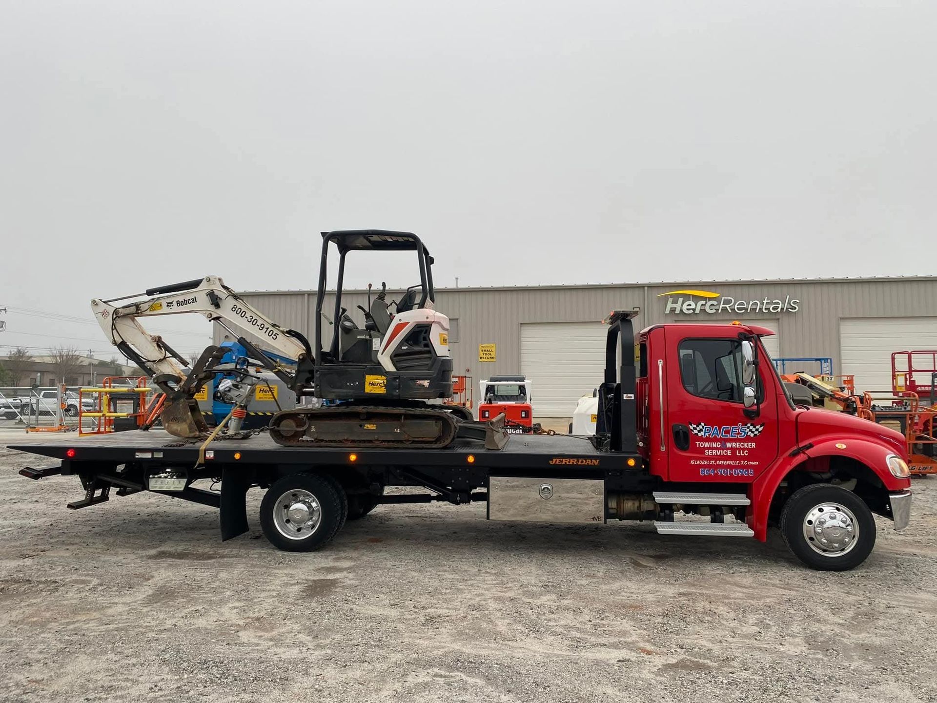 Red tow truck with a small excavator on its flatbed. Building in the background.