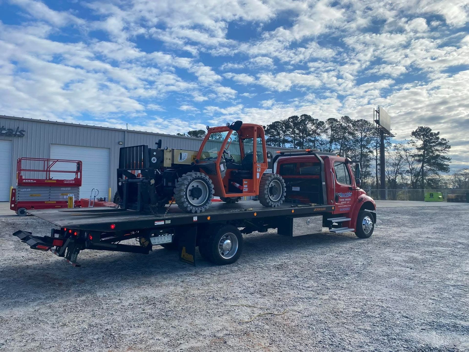 Orange forklift on a flatbed truck in front of a building under a cloudy sky.
