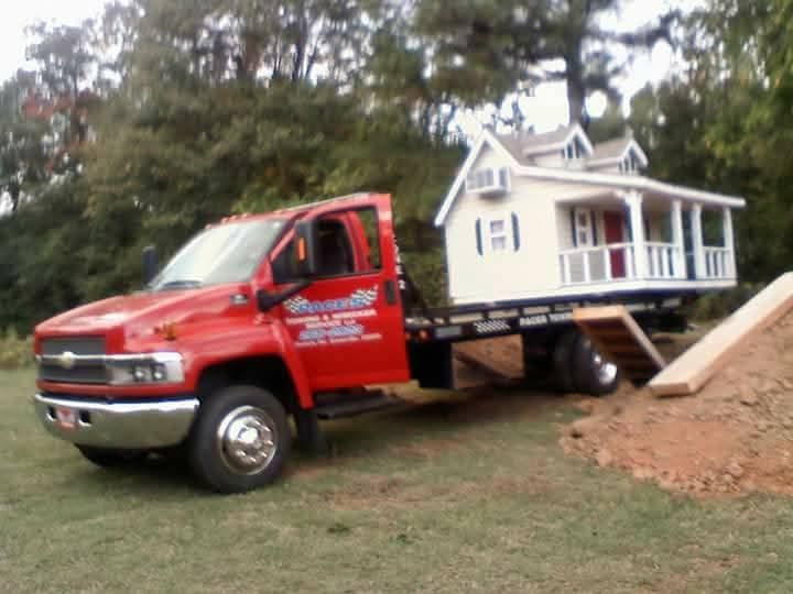 Red tow truck transporting a small white house with blue shutters on a flatbed trailer.