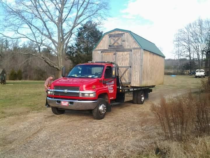 Red truck hauling a small wooden shed on a flatbed trailer in a grassy field.