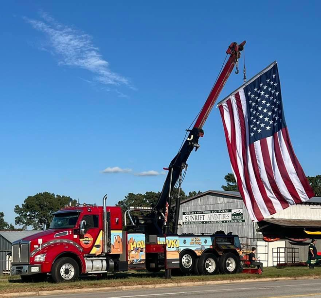 Red tow truck with raised crane beside a giant American flag on a trailer under a blue sky