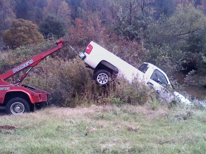 Red tow truck pulling a white pickup truck from a ditch along a grassy bank.