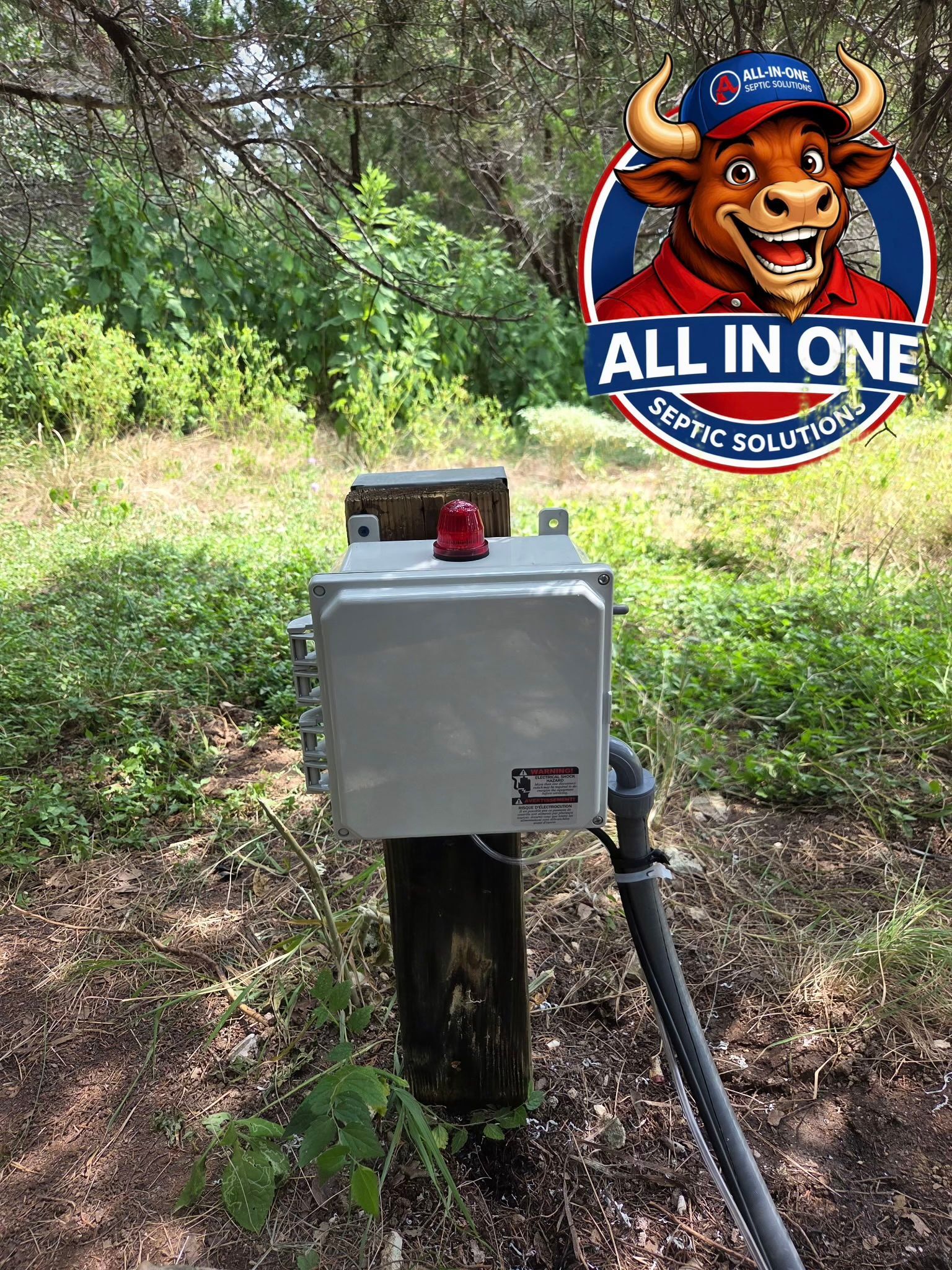 Gray electrical box on a wooden post in a wooded area, with a red light on top and hose attached