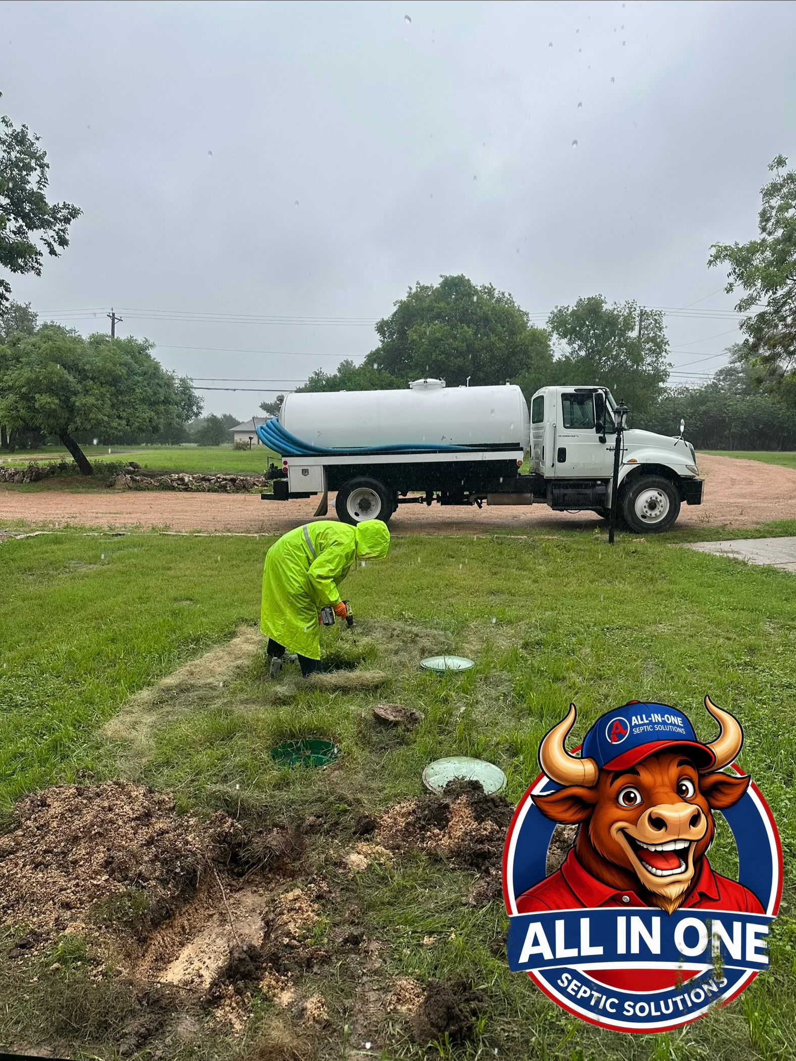 Worker in neon green crouches in a grassy field near a white tanker truck under cloudy skies.