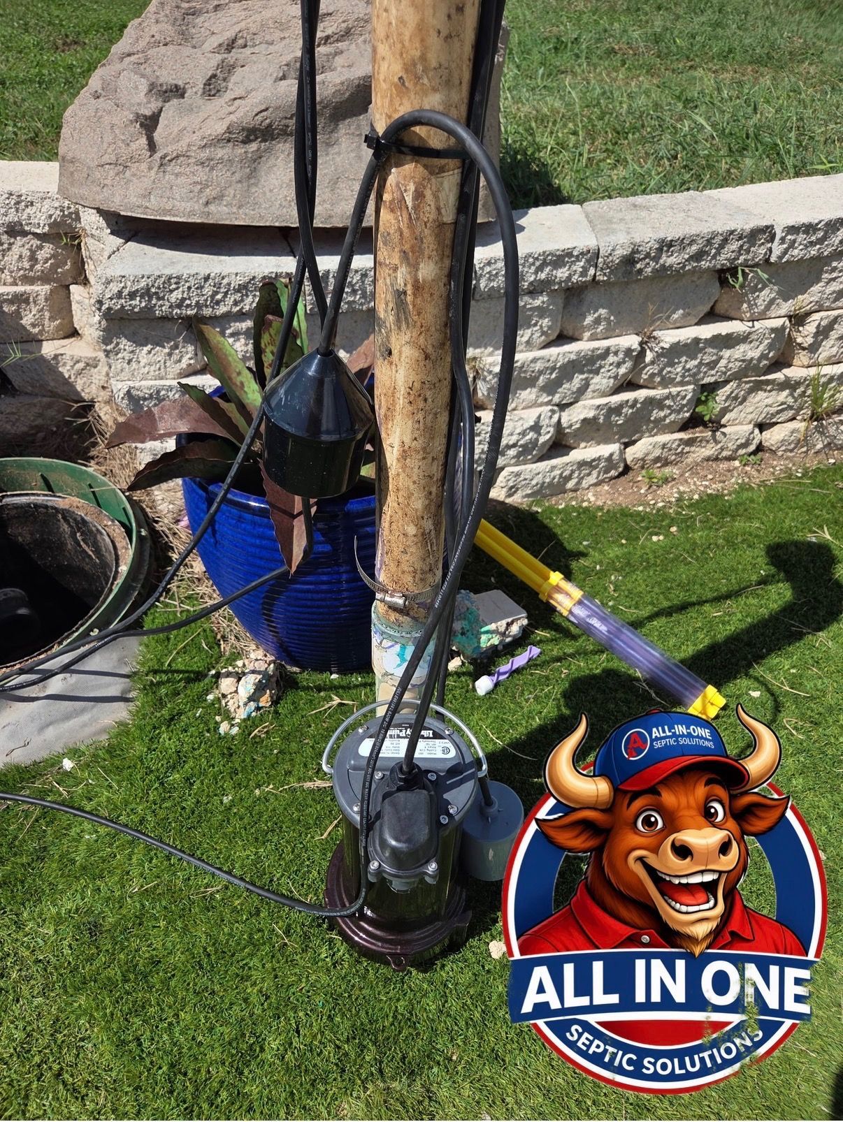 Blue pump with hoses strapped to a wooden post in a grassy yard beside a stone wall.