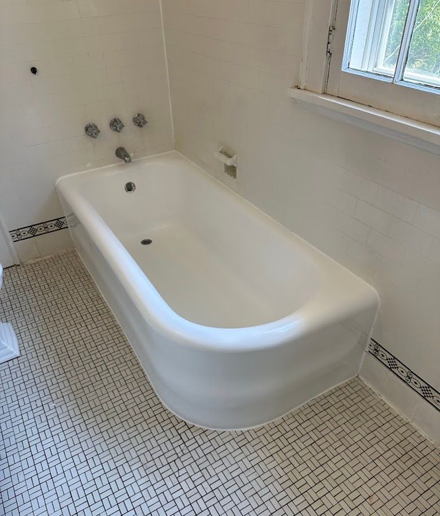 White bathtub in a tiled bathroom beside a window, with decorative mosaic floor border.