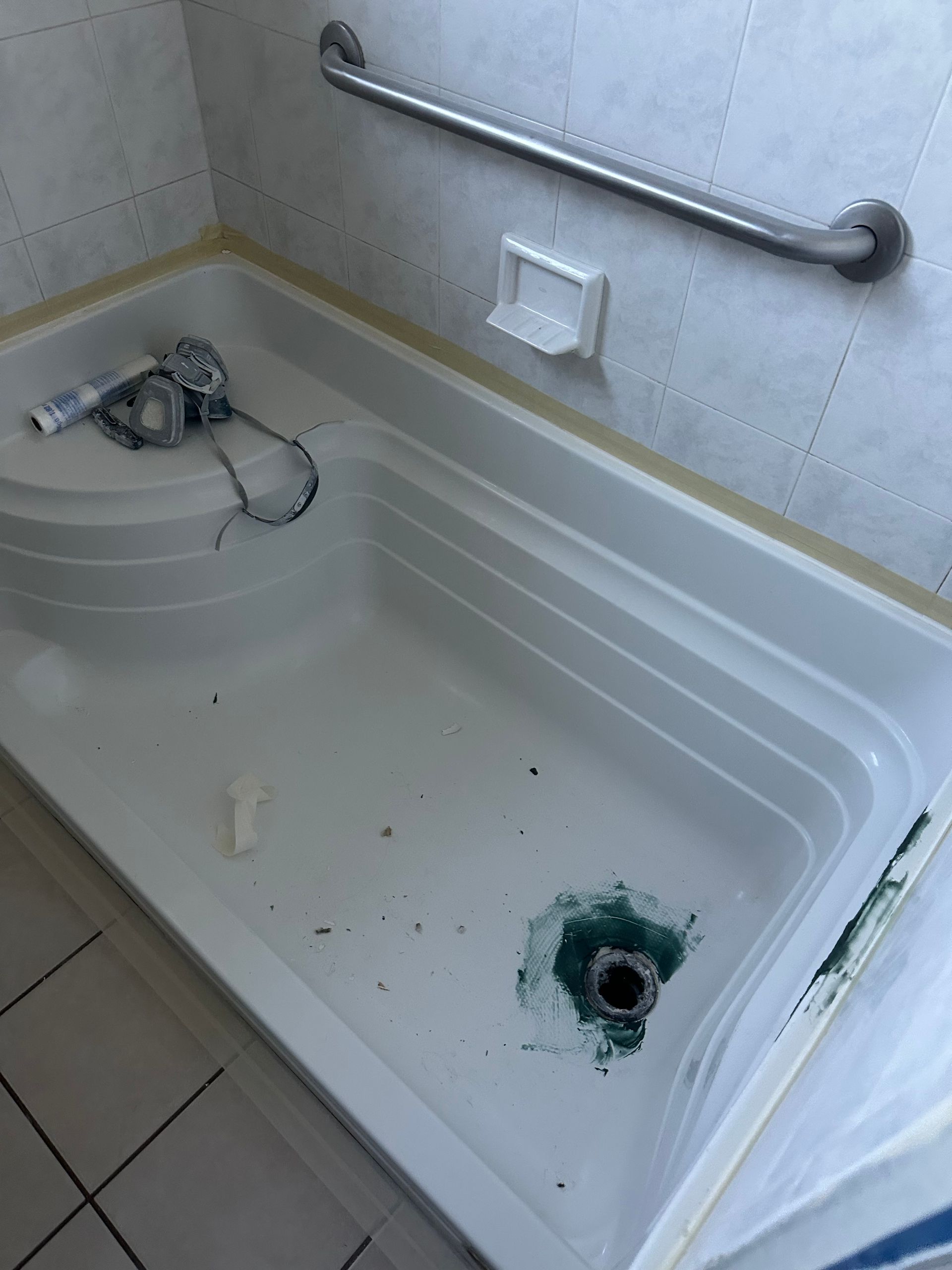 Bathtub with a damaged drain and debris, beside a shower grab bar and wall outlet.