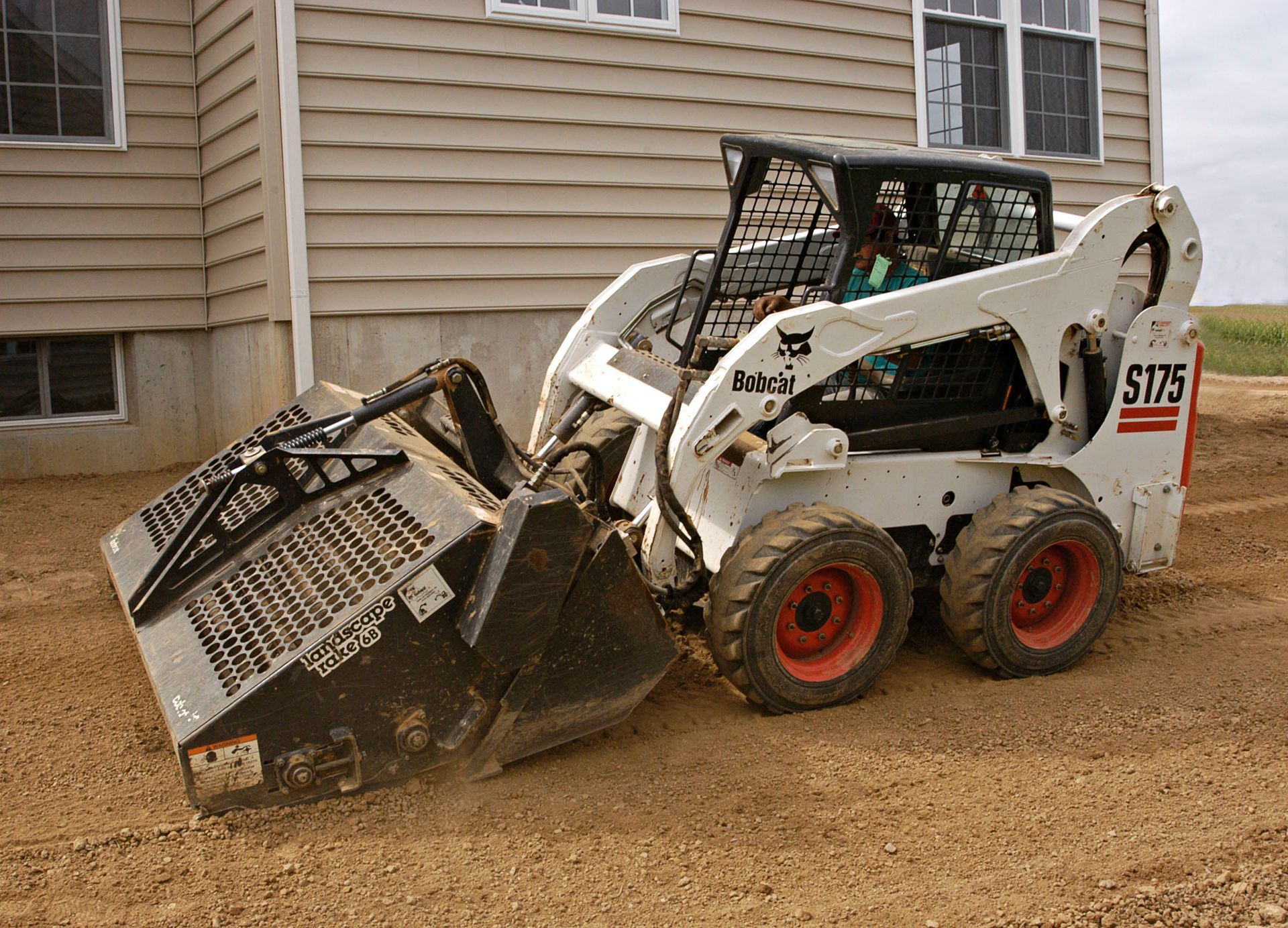 Bobcat S175 skid-steer loader scooping dirt near a light brown building. White with red wheels.